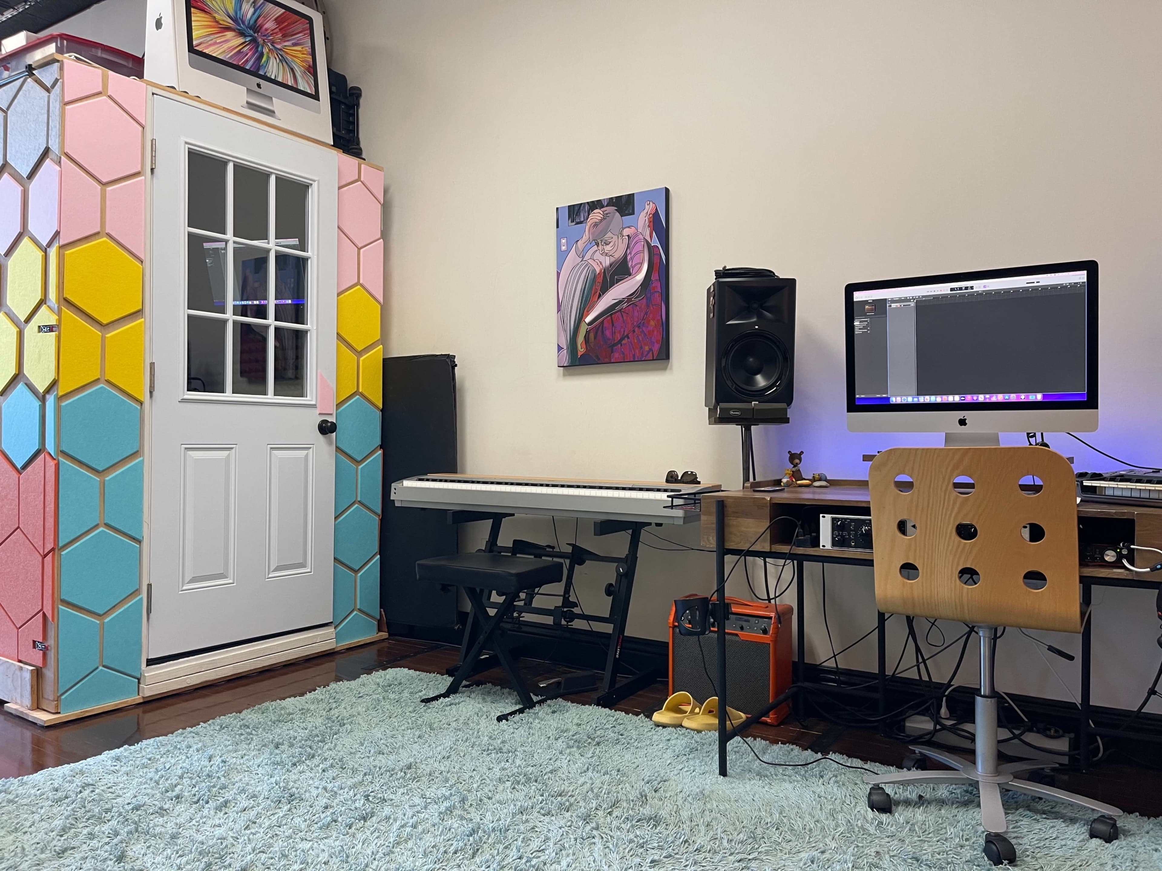 A music production studio featuring a keyboard, computer setup, and a colorful hexagonal wall near a door.