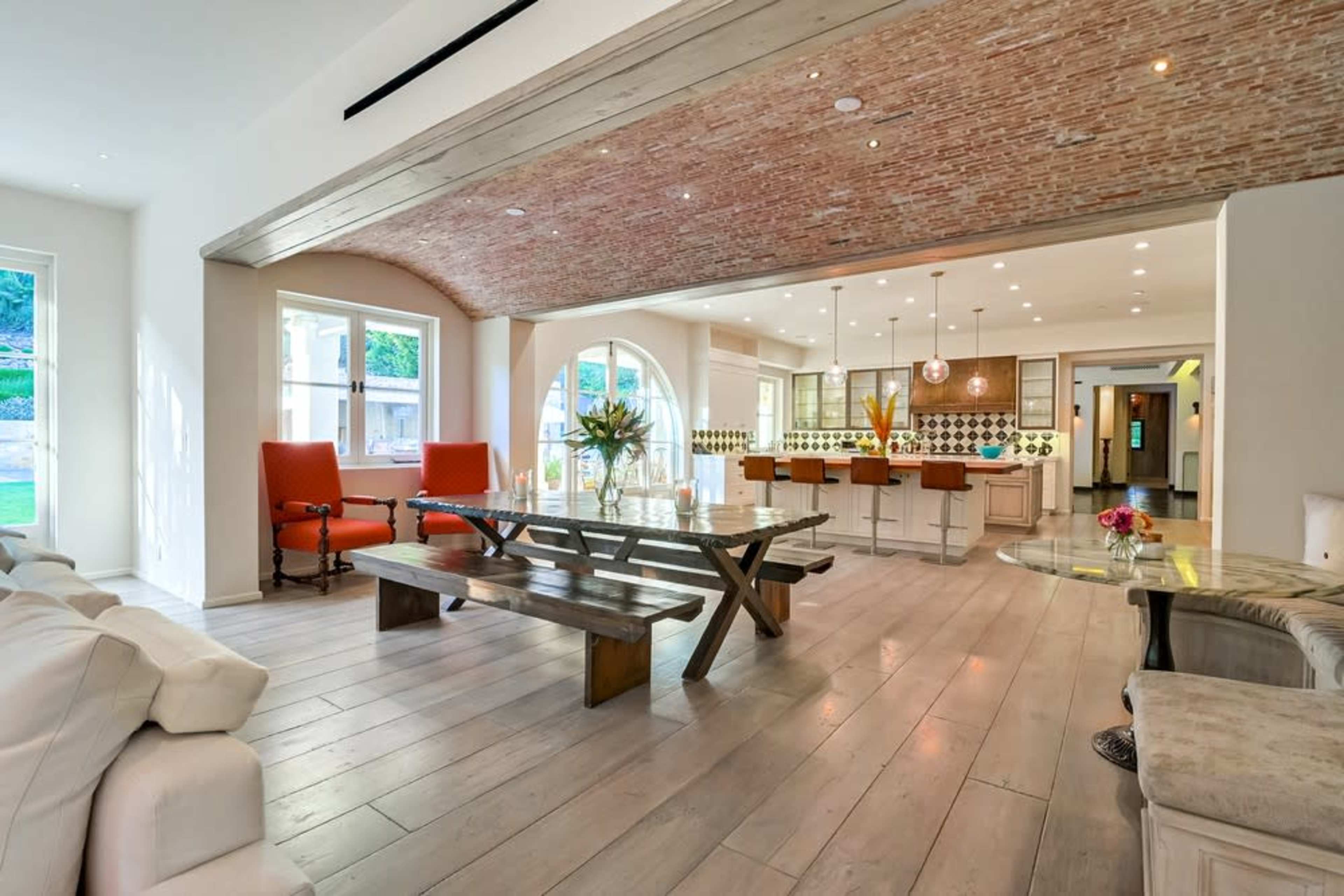A modern open-concept living space with a large wooden table, two orange chairs, and a kitchen area featuring a distinctive tile backsplash.