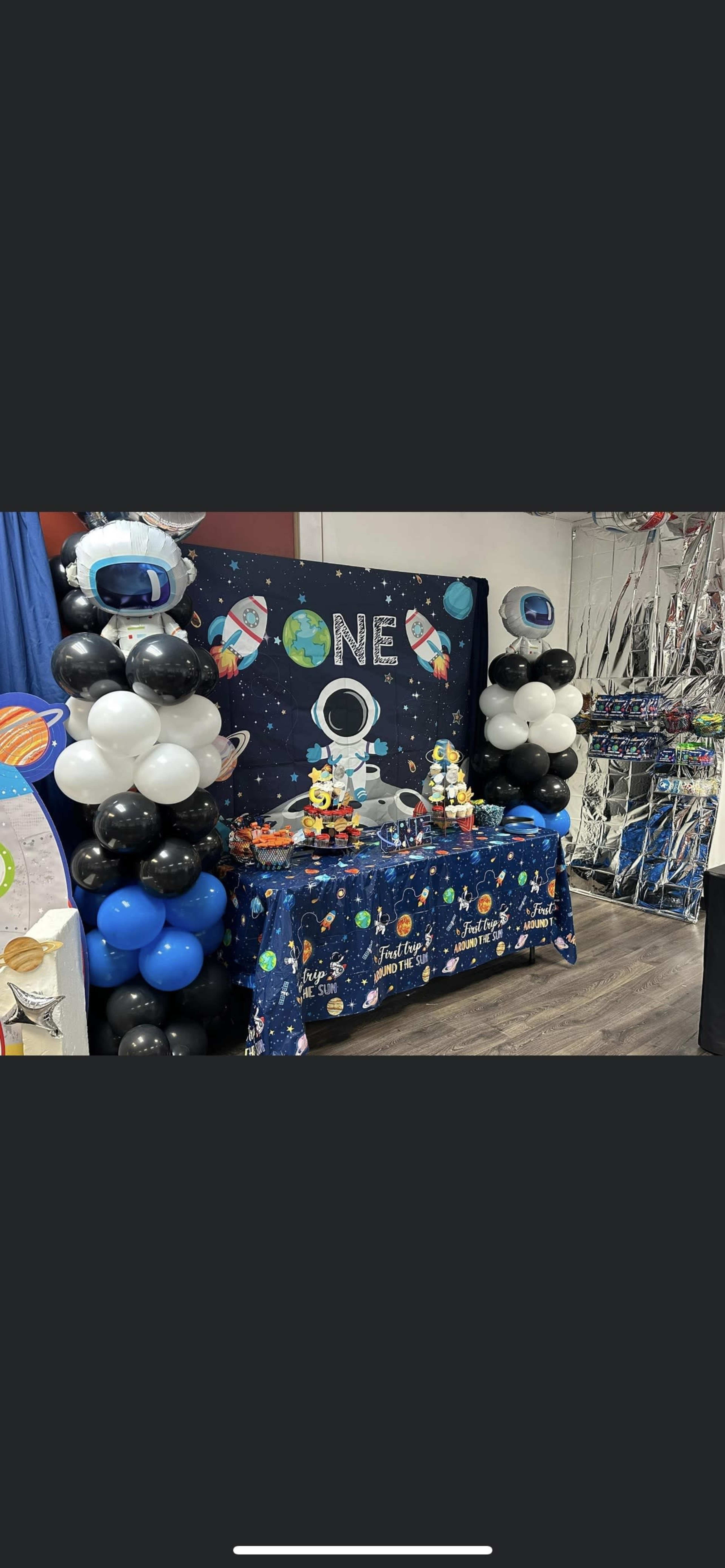 A birthday party setup features a space-themed table adorned with balloons and decorations, displaying a cake and treats under a backdrop that reads "ONE."