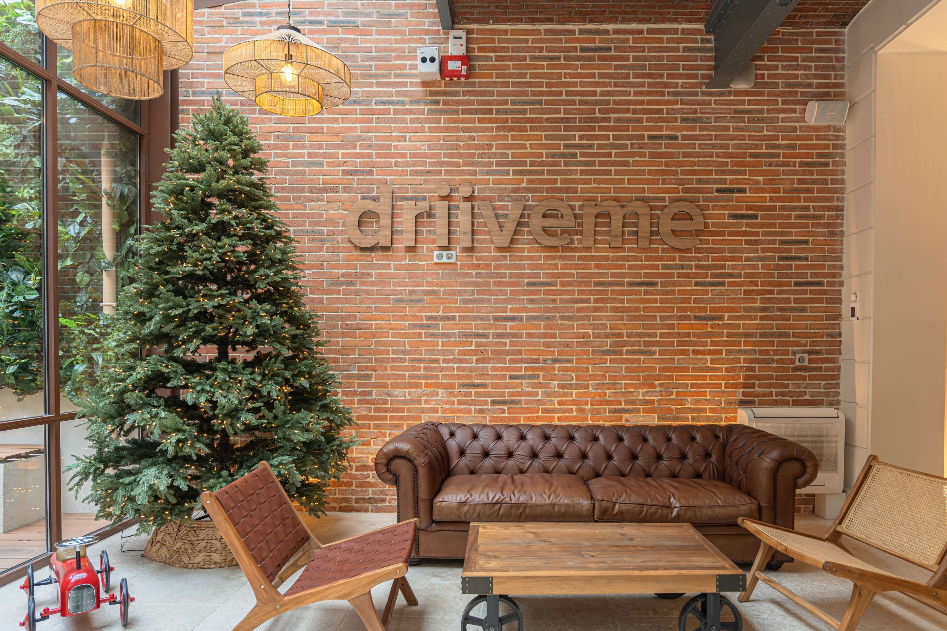 A cozy indoor space features a brick wall with the word "driveme," a large Christmas tree, a brown leather sofa, and wooden chairs around a coffee table.