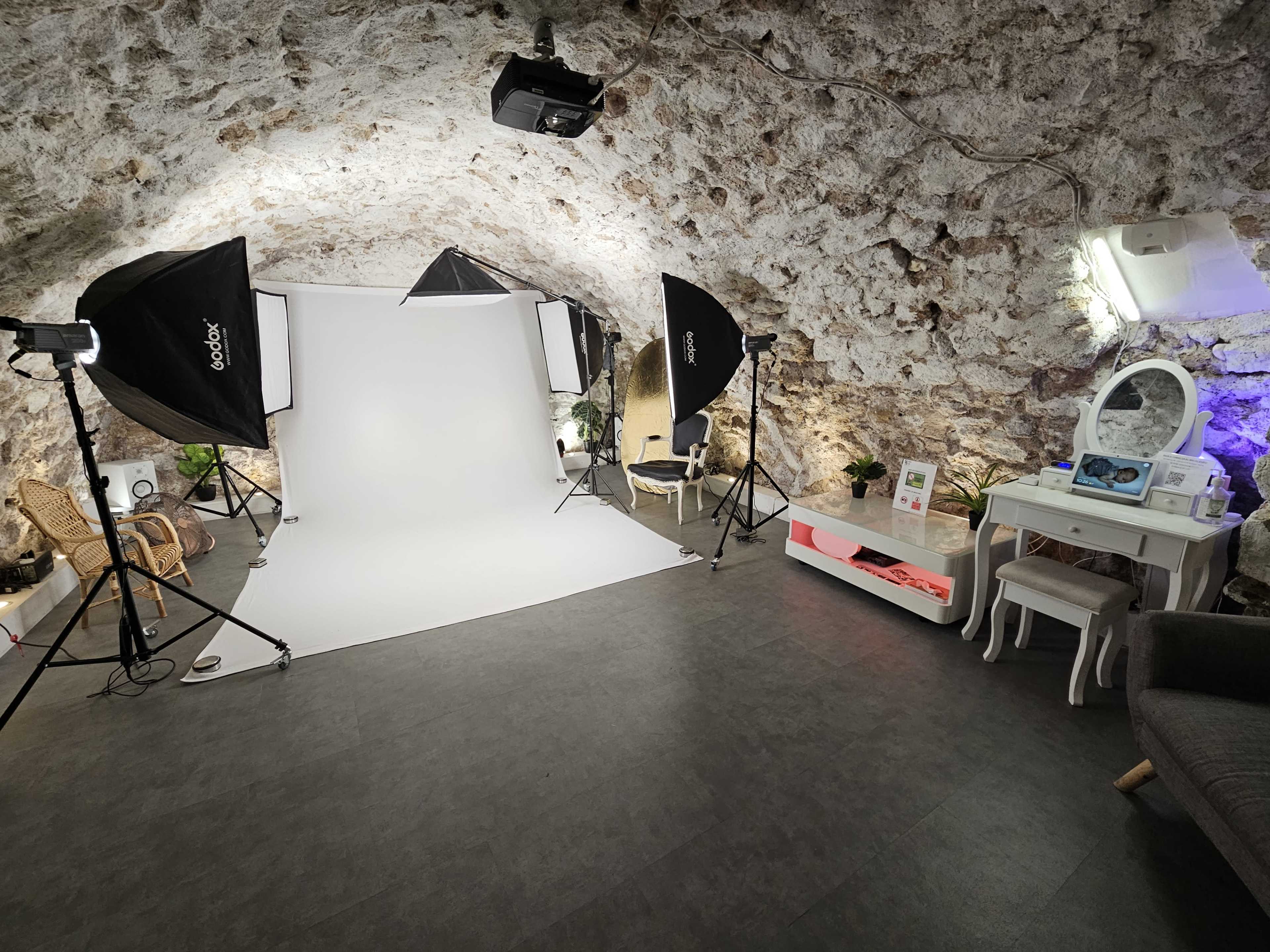 A photography studio set up in a stone-walled room, featuring lighting equipment, a white backdrop, and a vanity table.