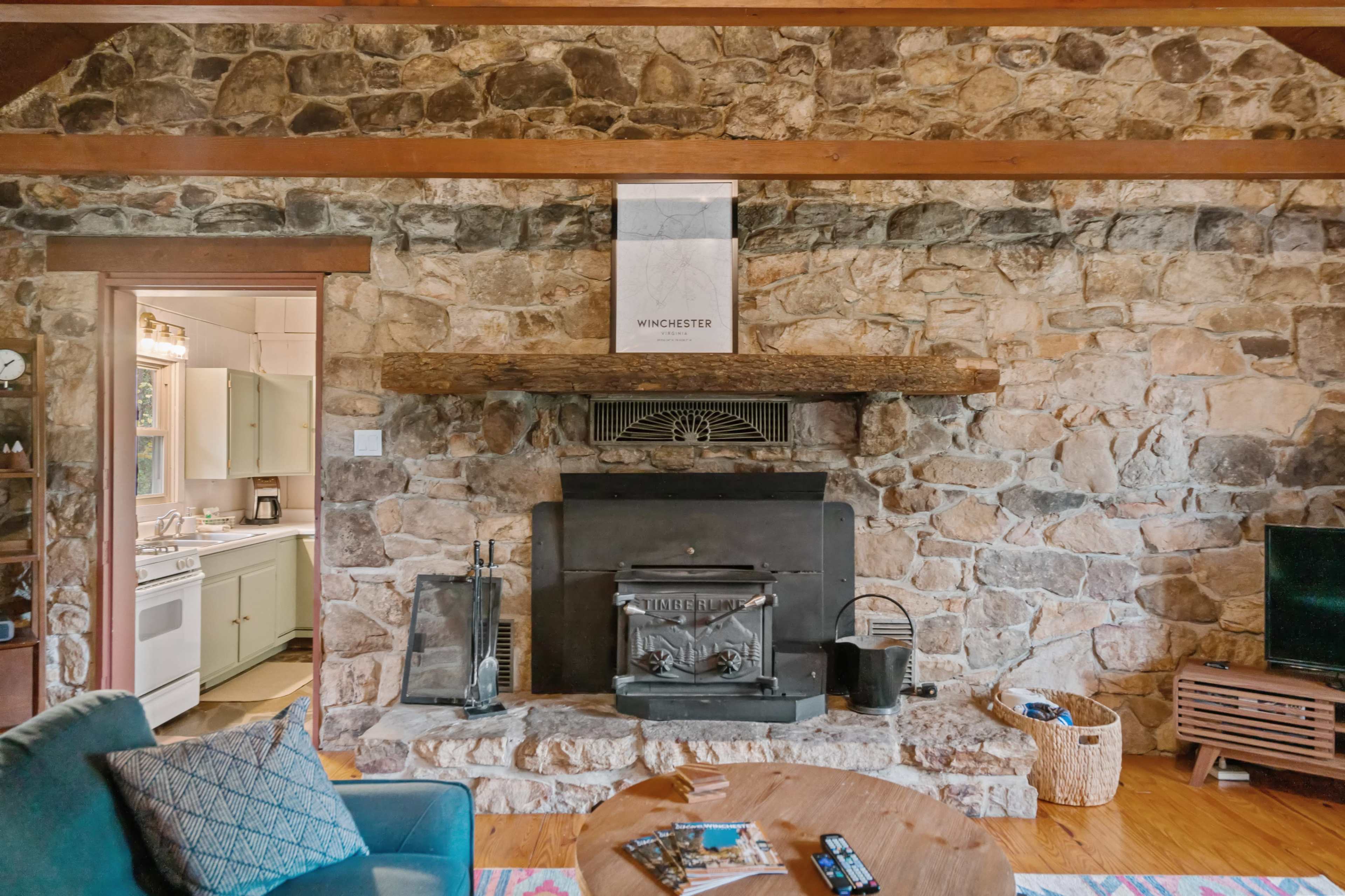 A living room with a stone wall, featuring a central fireplace with a wooden mantel, surrounded by a blue armchair and a coffee table.