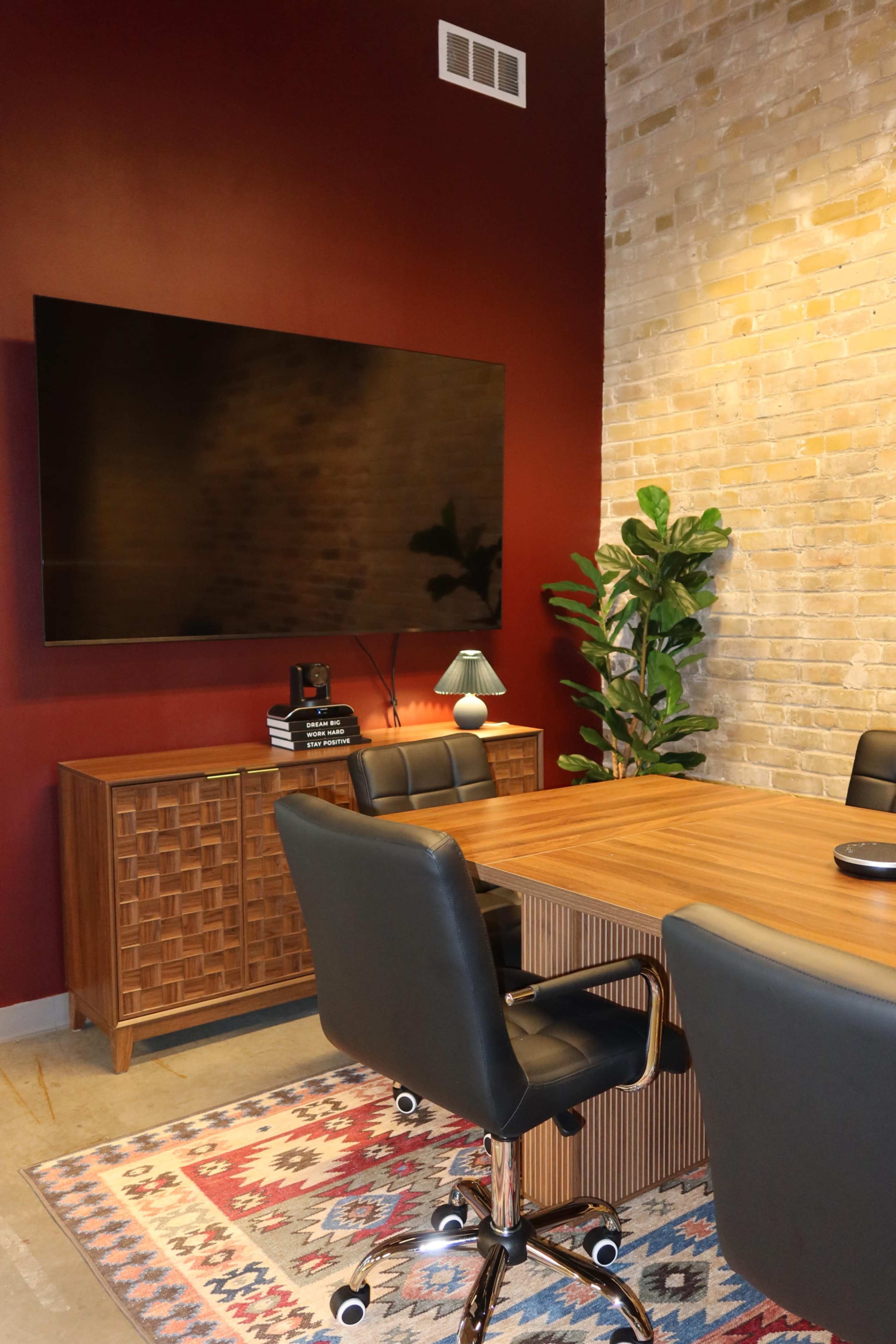 The image shows a modern conference room with a wooden table, black chairs, a large wall-mounted TV, and decorative elements including a lamp and a potted plant against a red and exposed brick wall.