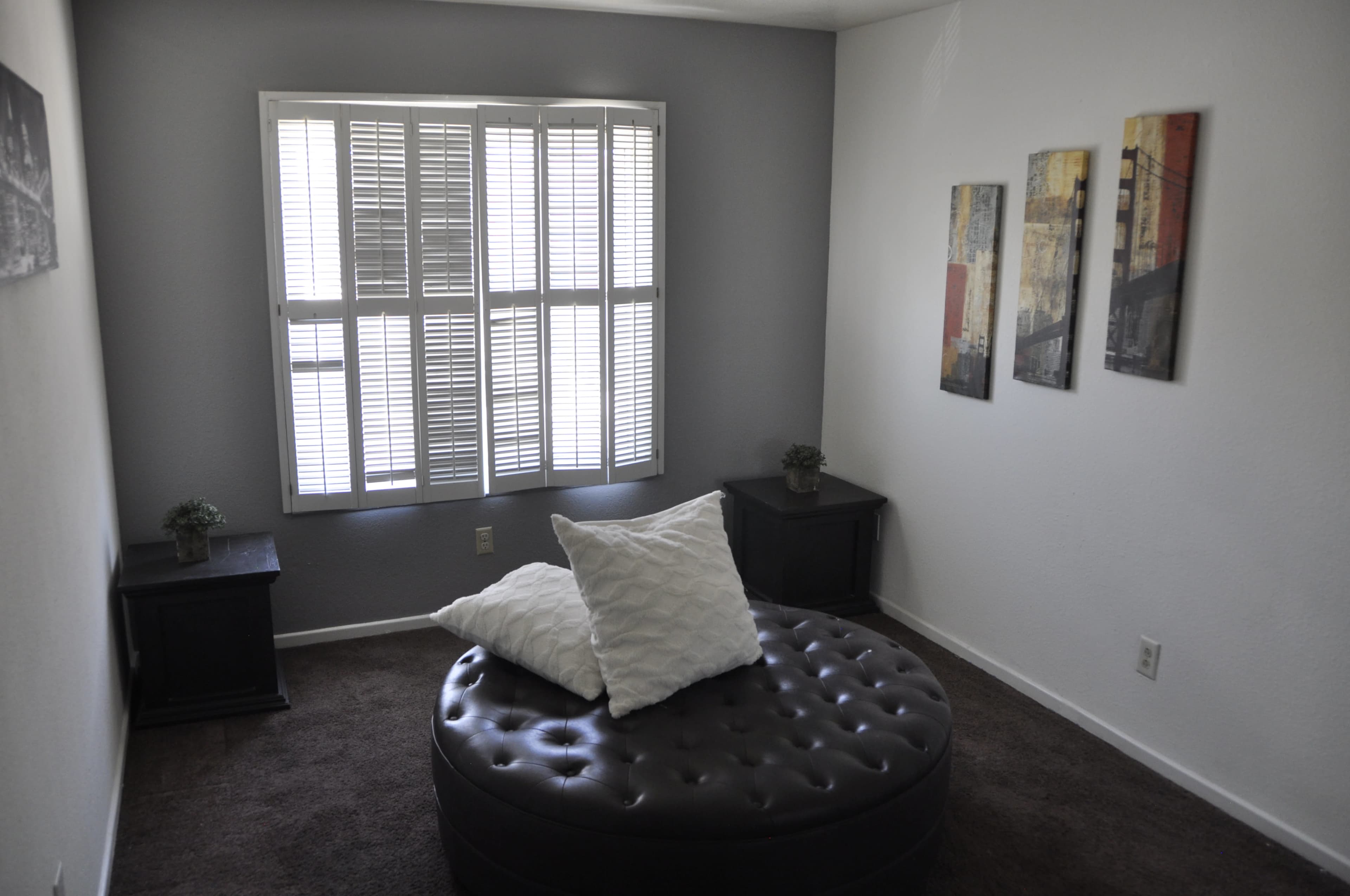 The image depicts a small, furnished living space featuring a circular upholstered ottoman, two side tables, and framed artwork on the wall, with a window providing natural light.