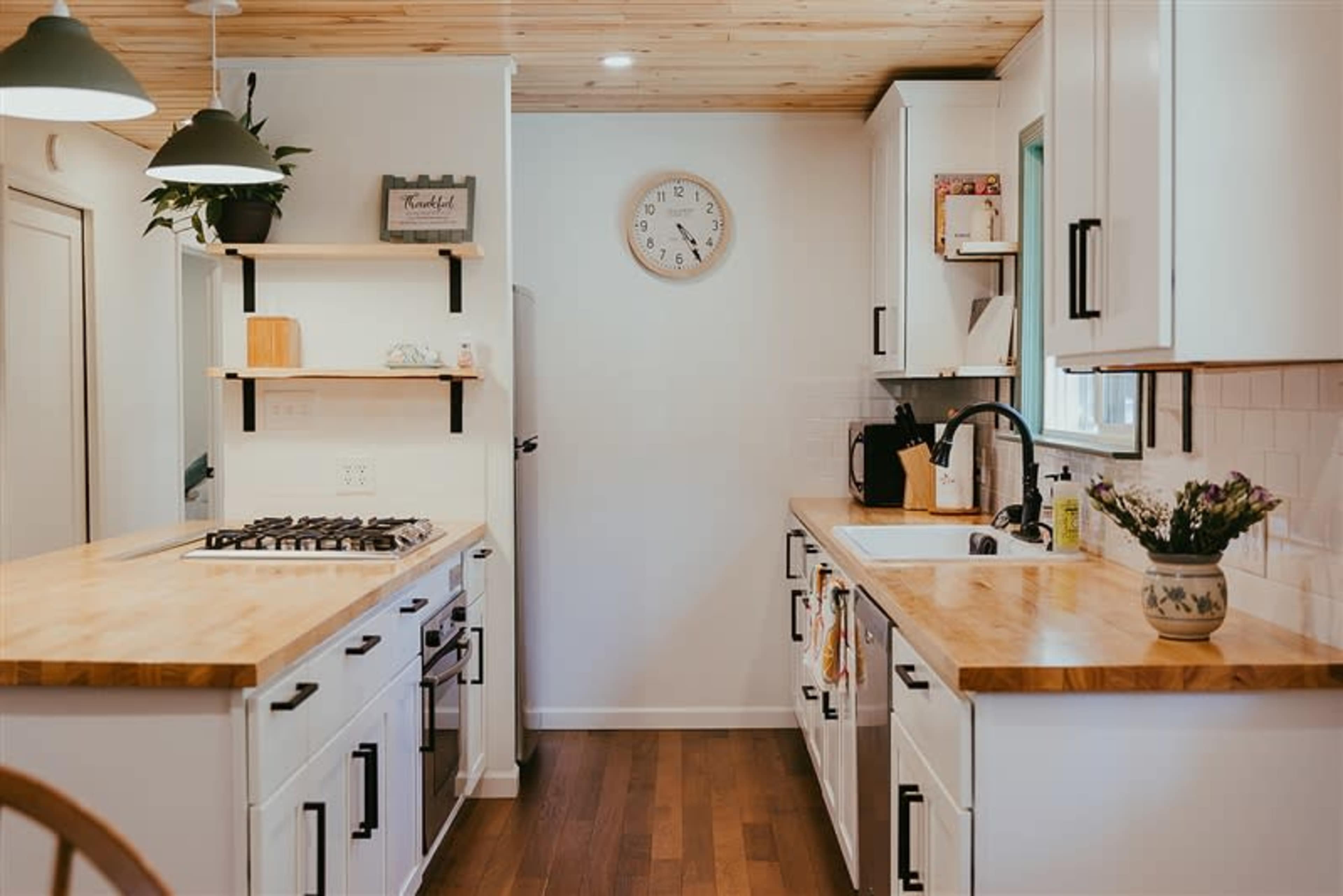 A bright kitchen features wooden countertops, white cabinetry, a clock on the wall, and open shelving with decorative items and plants.