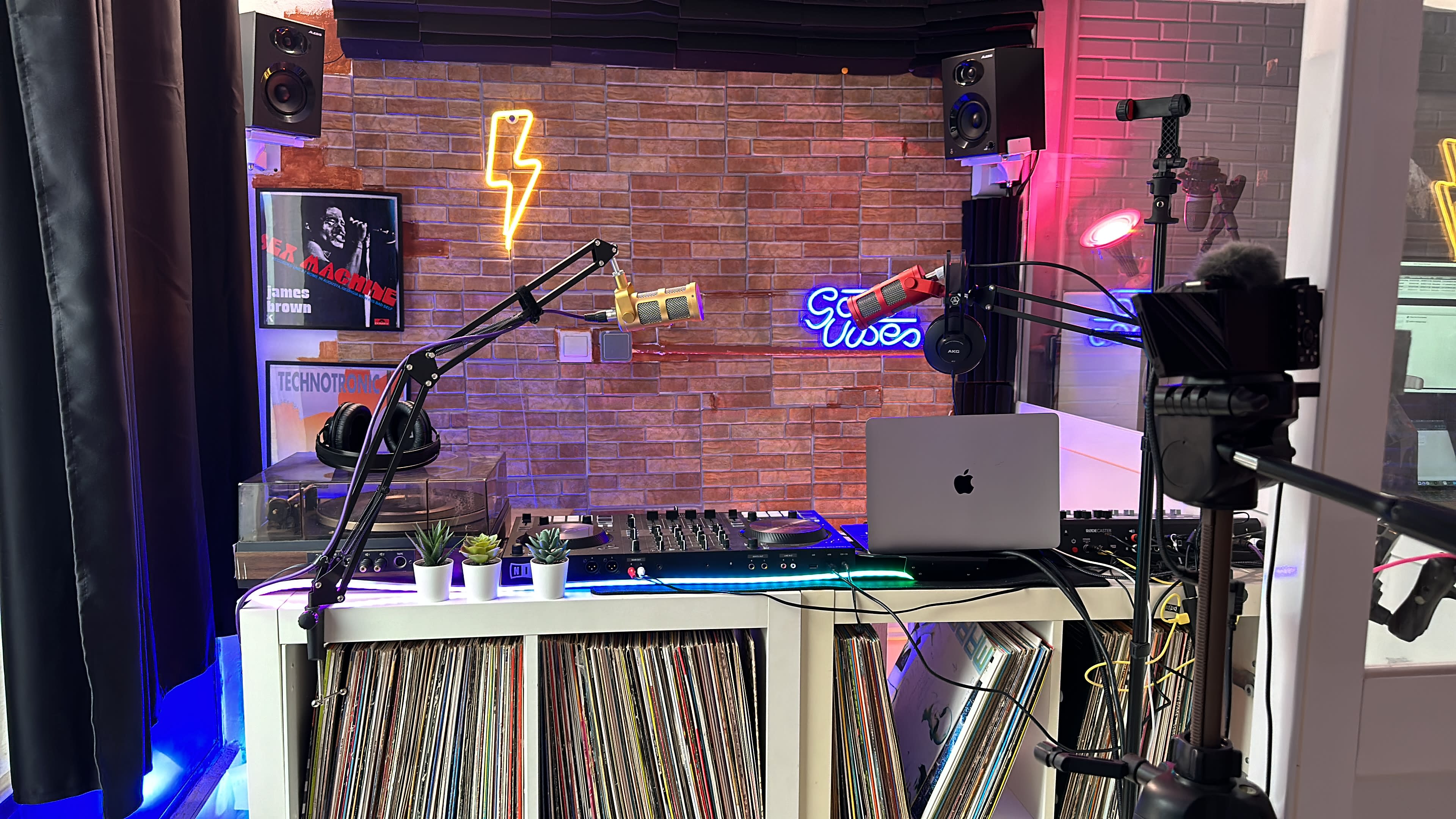 The image shows a DJ setup with a mixer, vinyl records, and various microphones, surrounded by a colorful backdrop featuring neon signs and studio equipment.
