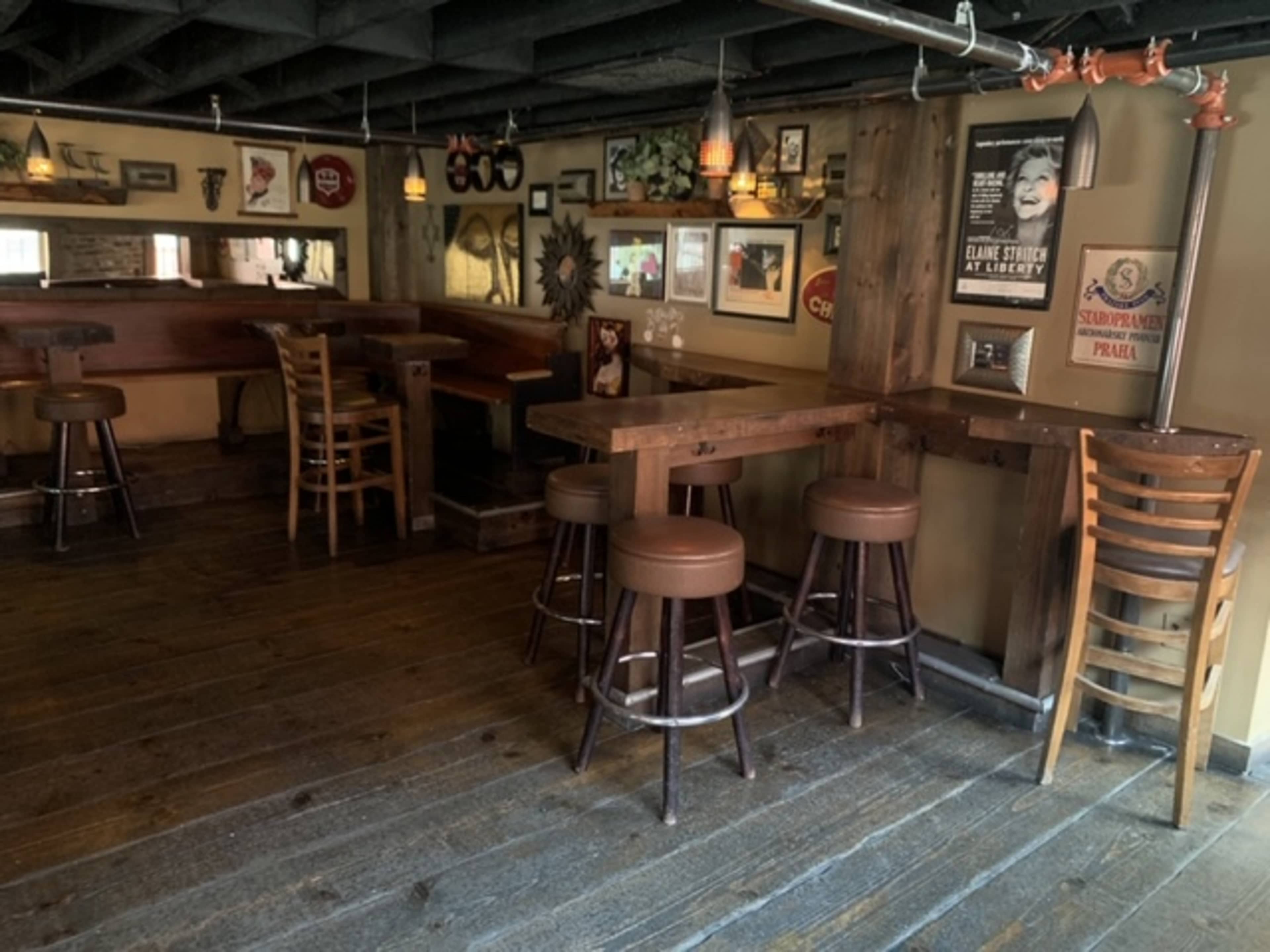 A rustic bar interior with wooden floors, a few empty high-top tables, and bar stools under dim lighting.
