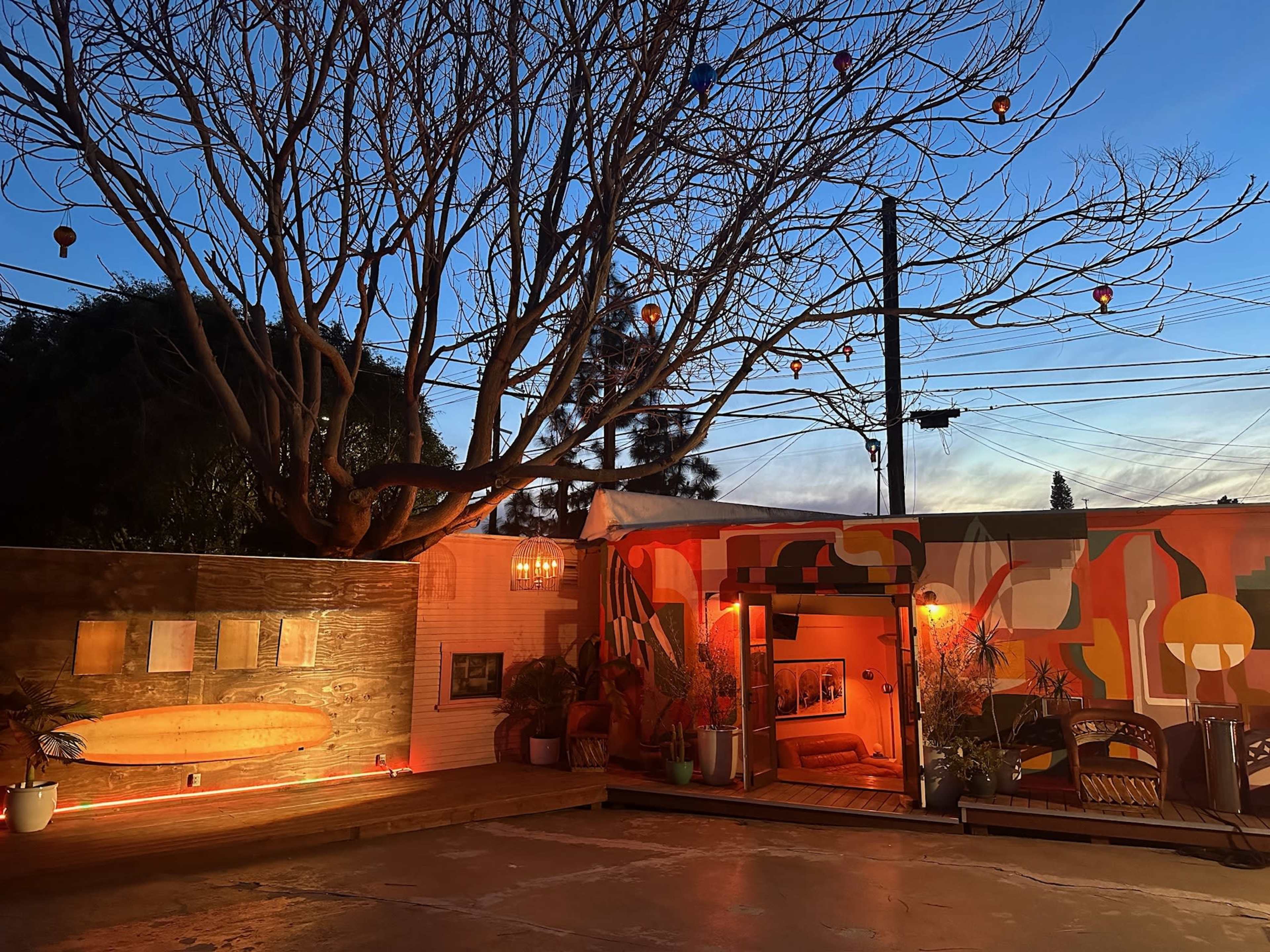 A colorful mural and a wooden surfboard adorn a patio area under a tree, with decorative lights hanging overhead as twilight approaches.