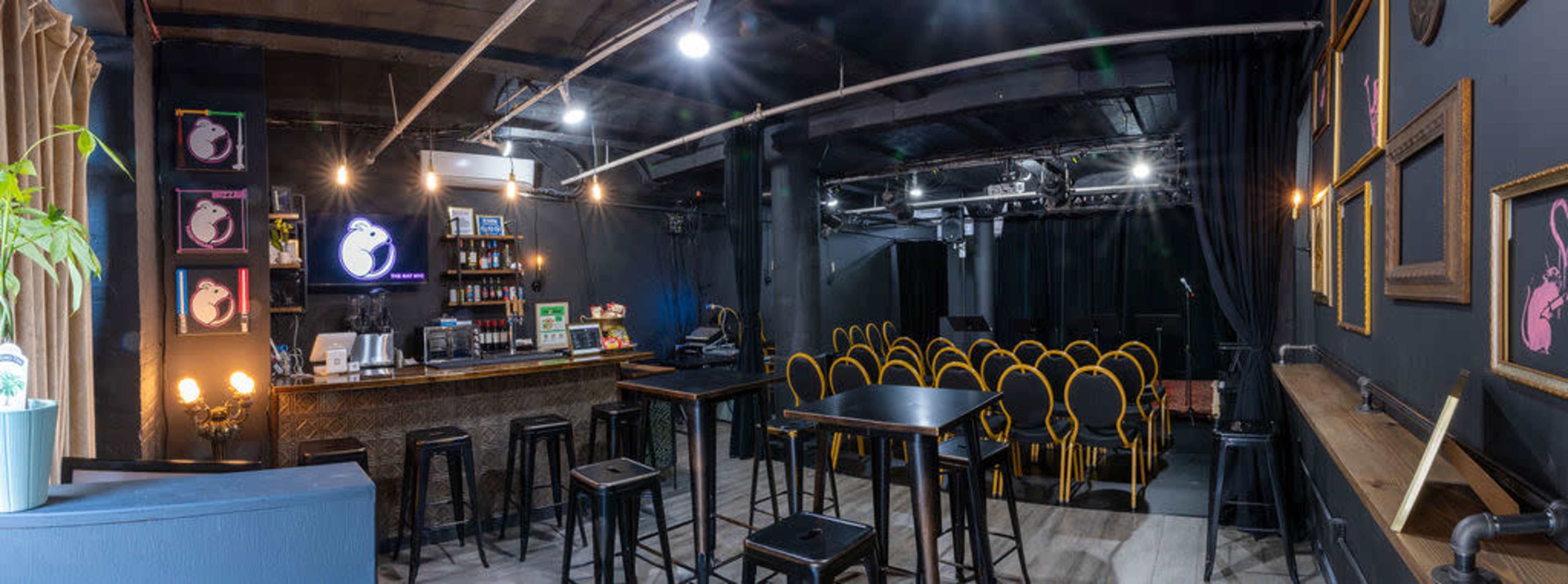 A dimly lit bar area with high tables and stools, a counter with drinks, and a small stage in the background surrounded by rows of yellow chairs.