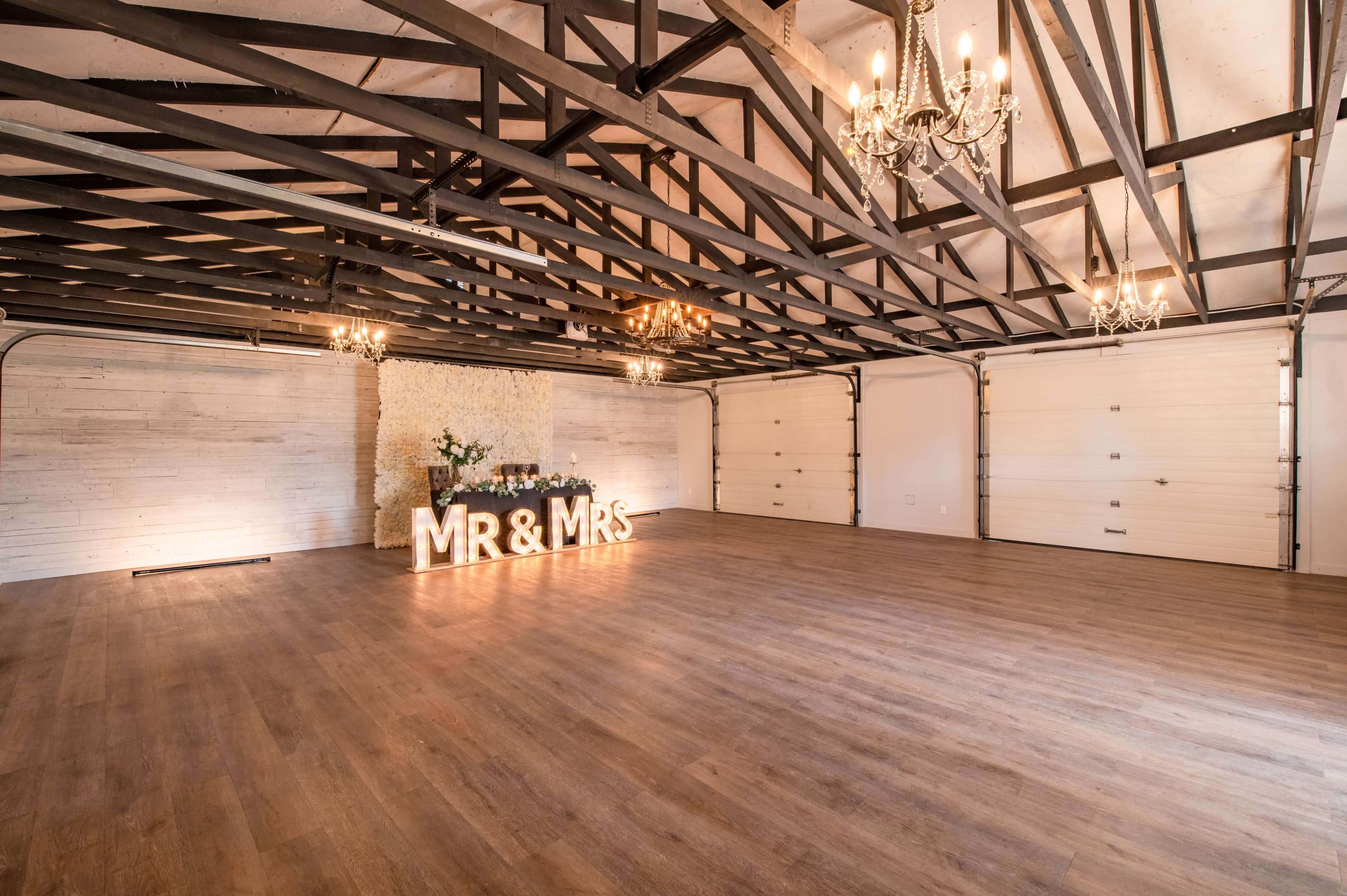 The image shows an empty event space with wooden flooring, exposed beams, chandeliers, and a decorative backdrop featuring "MR & MRS" illuminated in lights.