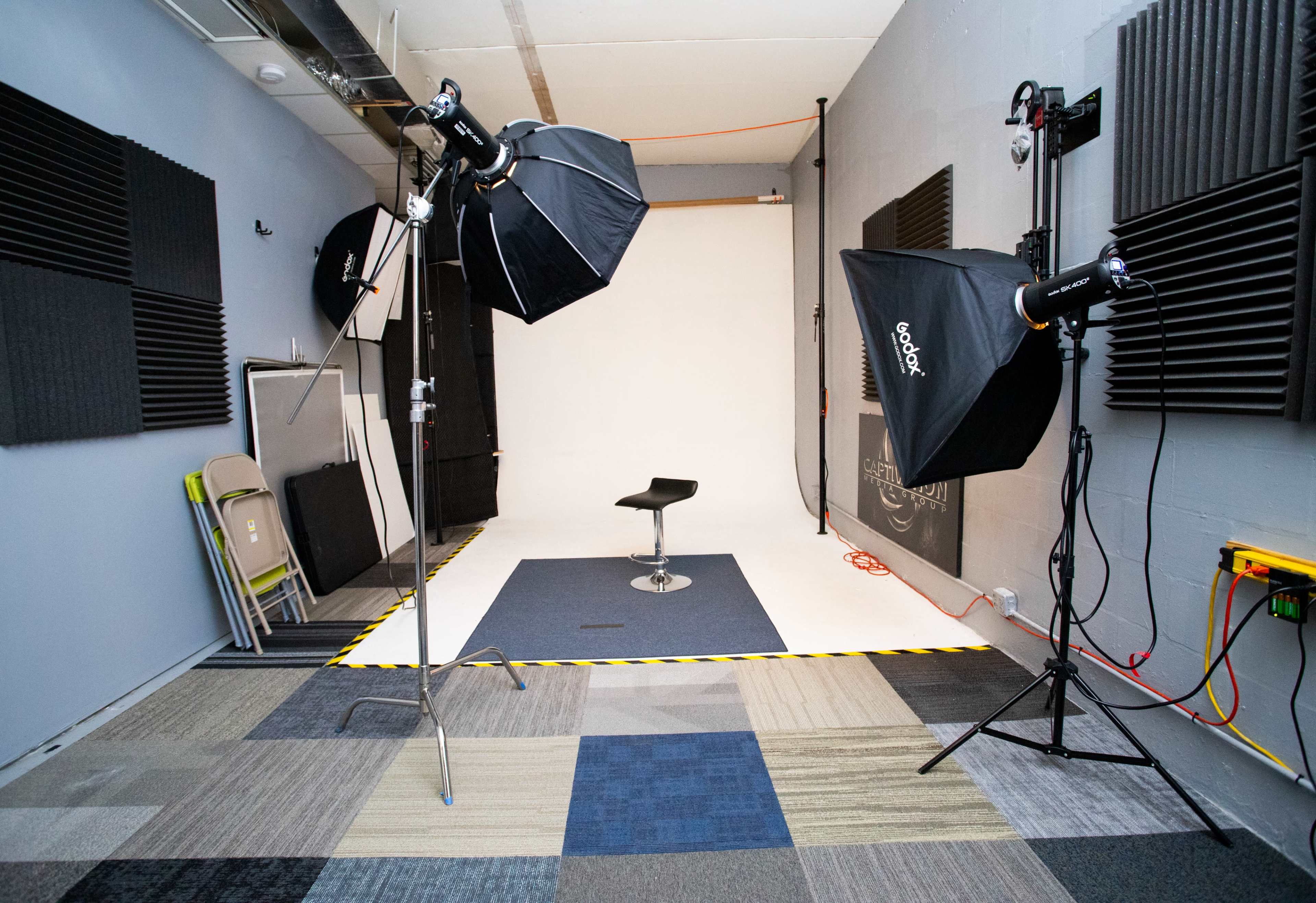 The image shows a photography studio with softbox lighting, a black chair positioned on a dark mat, and soundproofing panels along the walls.