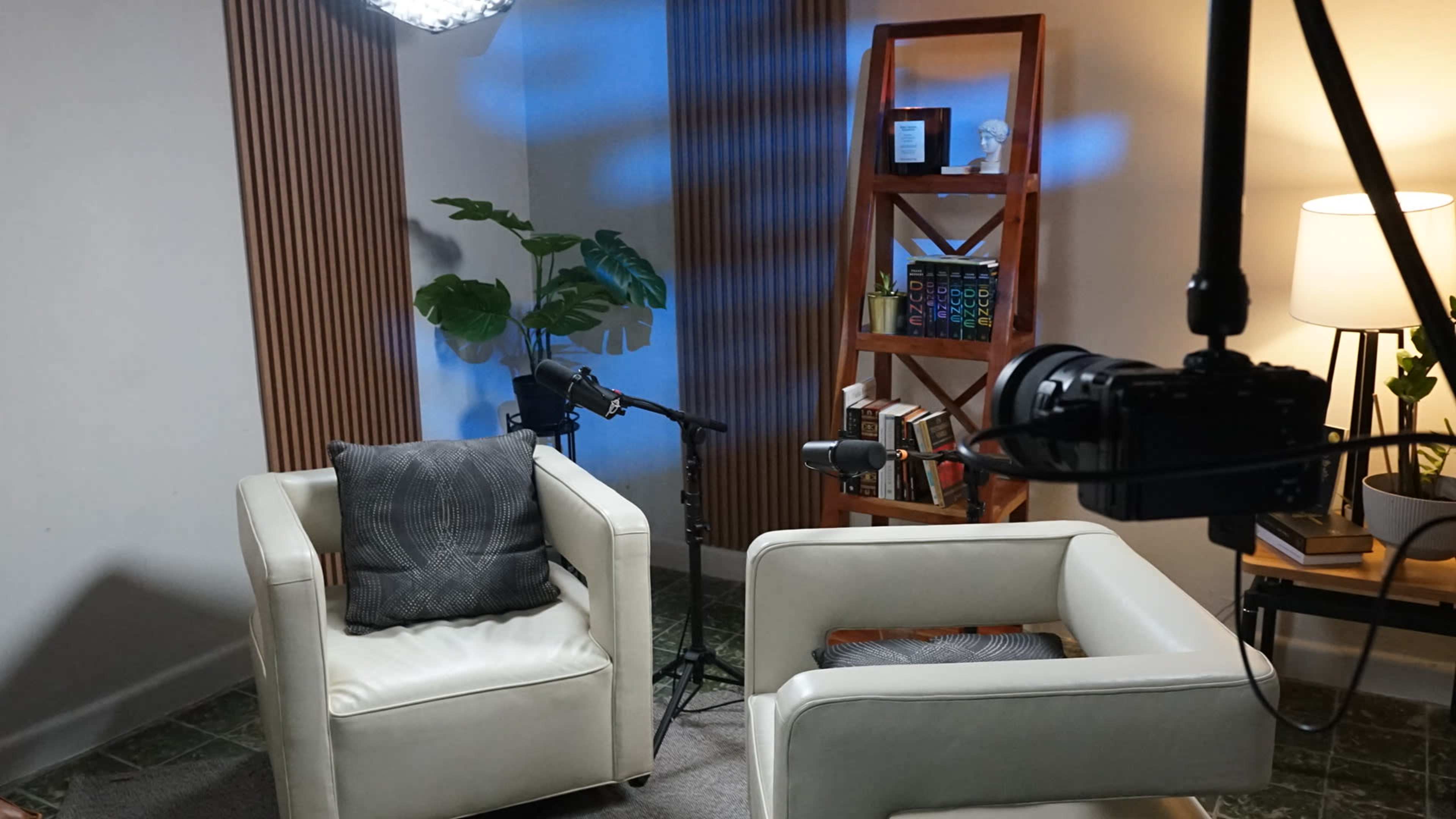 A well-lit interview space features two white chairs, microphones set up, and a bookshelf filled with books and decorative items in the background.