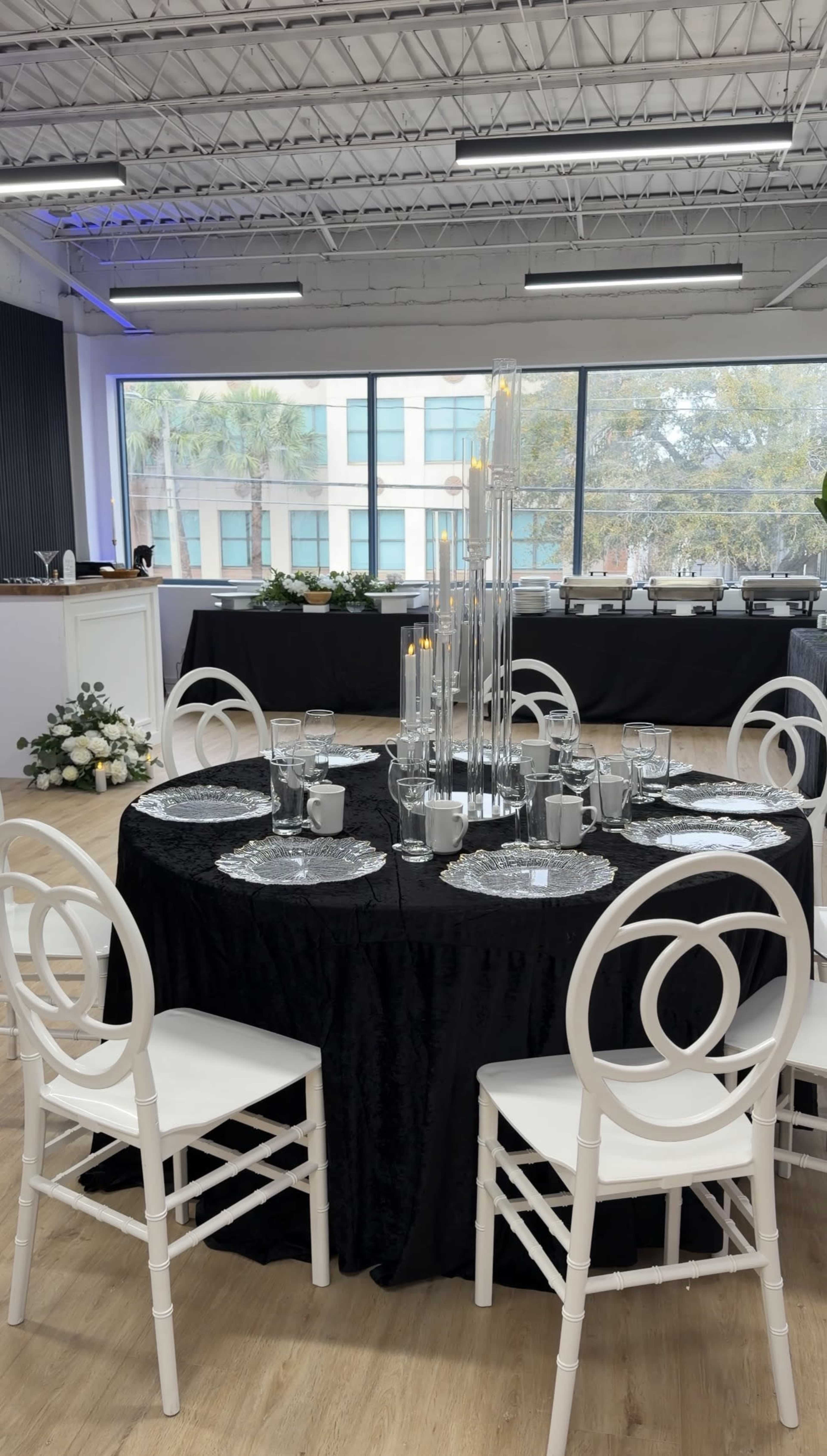 A round black table is set with elegant glassware and silver accents, surrounded by white chairs in a spacious room with large windows overlooking greenery.