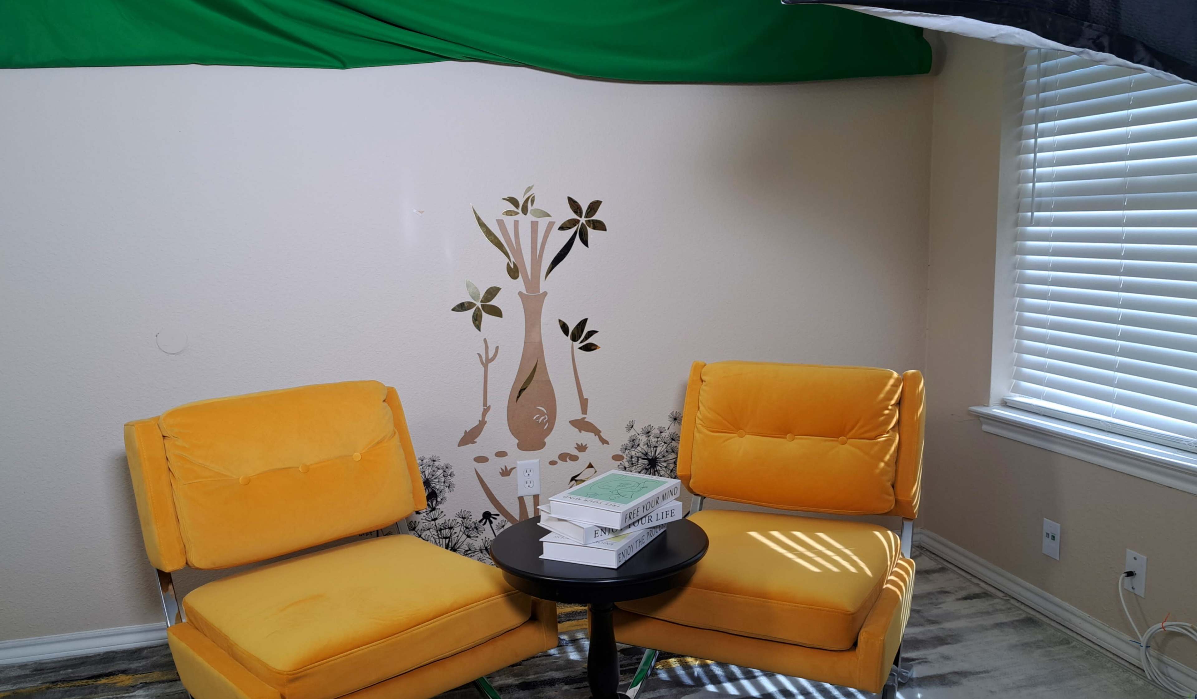 The image shows a small room with two yellow armchairs facing a black table, featuring a wall decoration of a tree and several books stacked on the table.