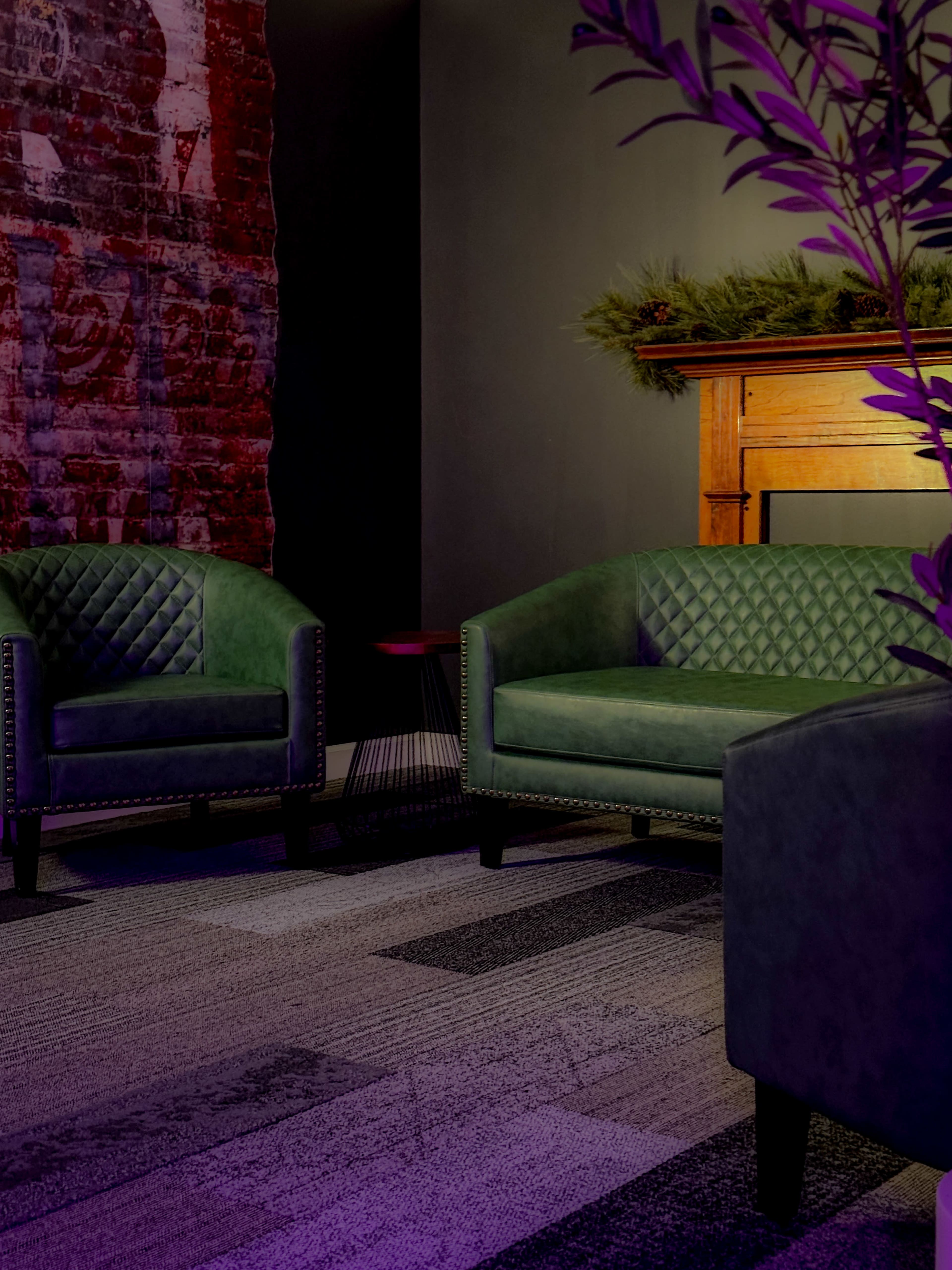 The image features two green velvet armchairs with a textured pattern, positioned against a brick wall and near a wooden mantel.
