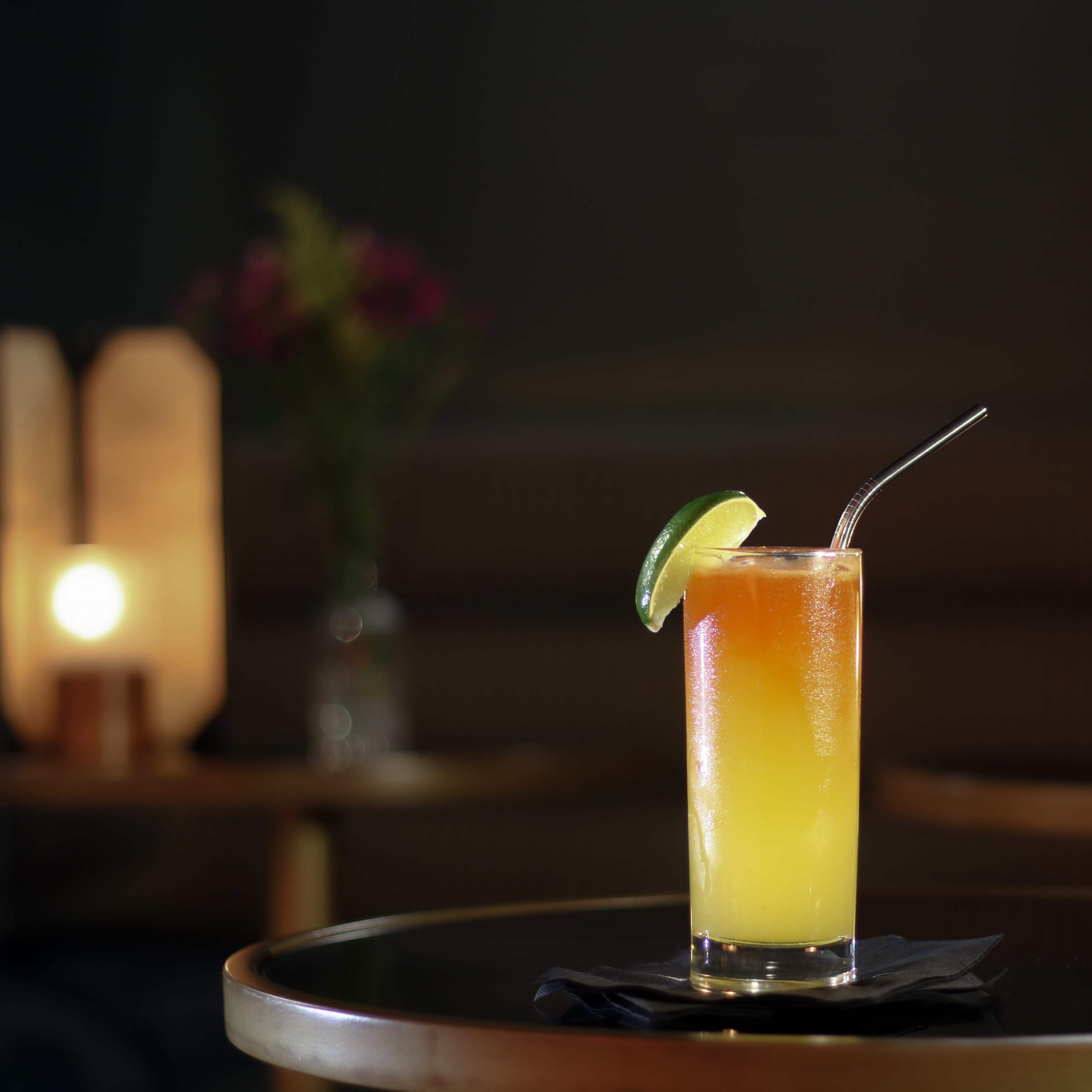 A tall glass of a gradient-colored cocktail sits on a table with a lime wedge and a straw, illuminated softly by ambient lighting.