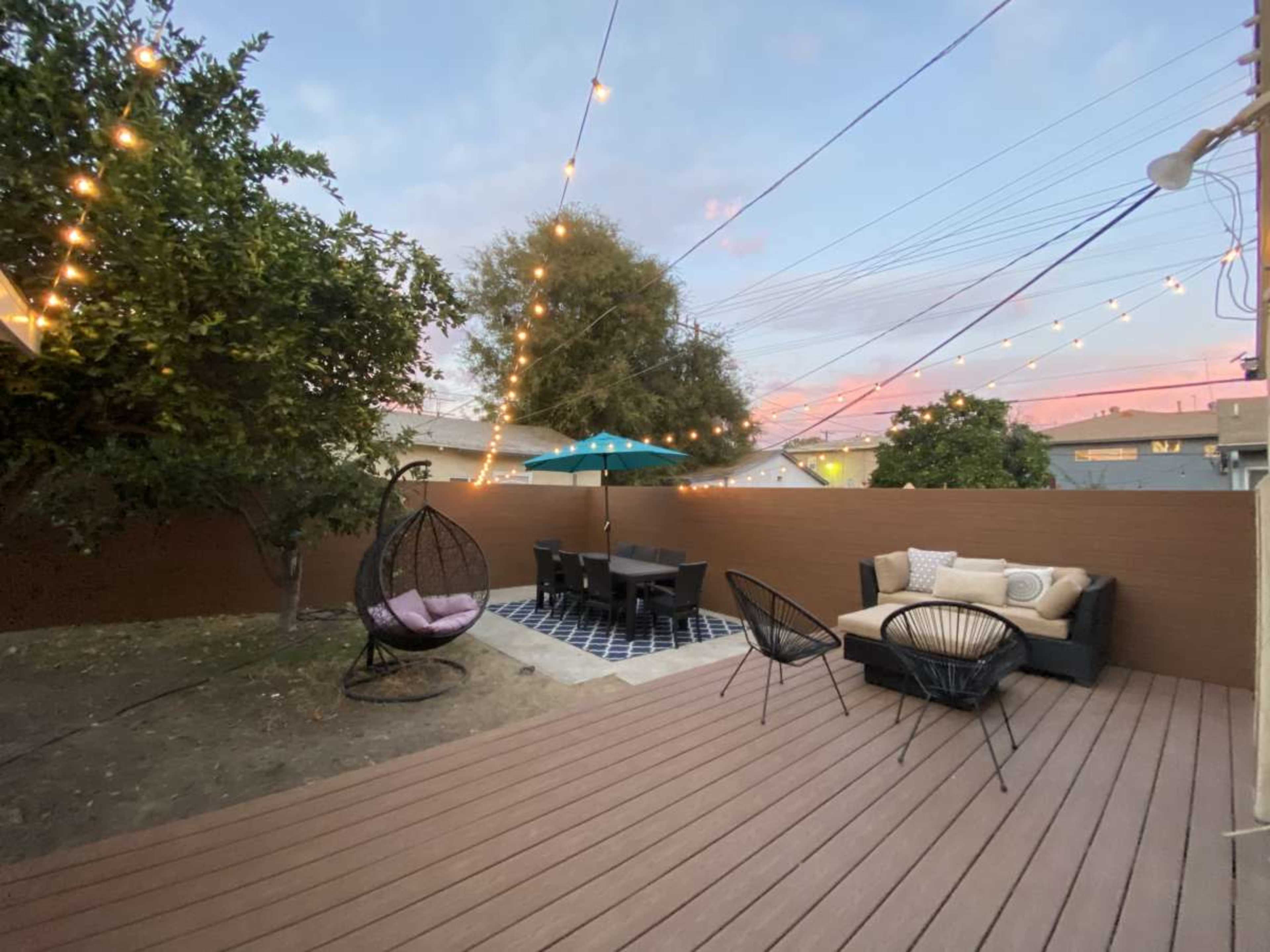 The image shows a backyard patio with a wooden deck, outdoor seating, a table under an umbrella, and string lights overhead.