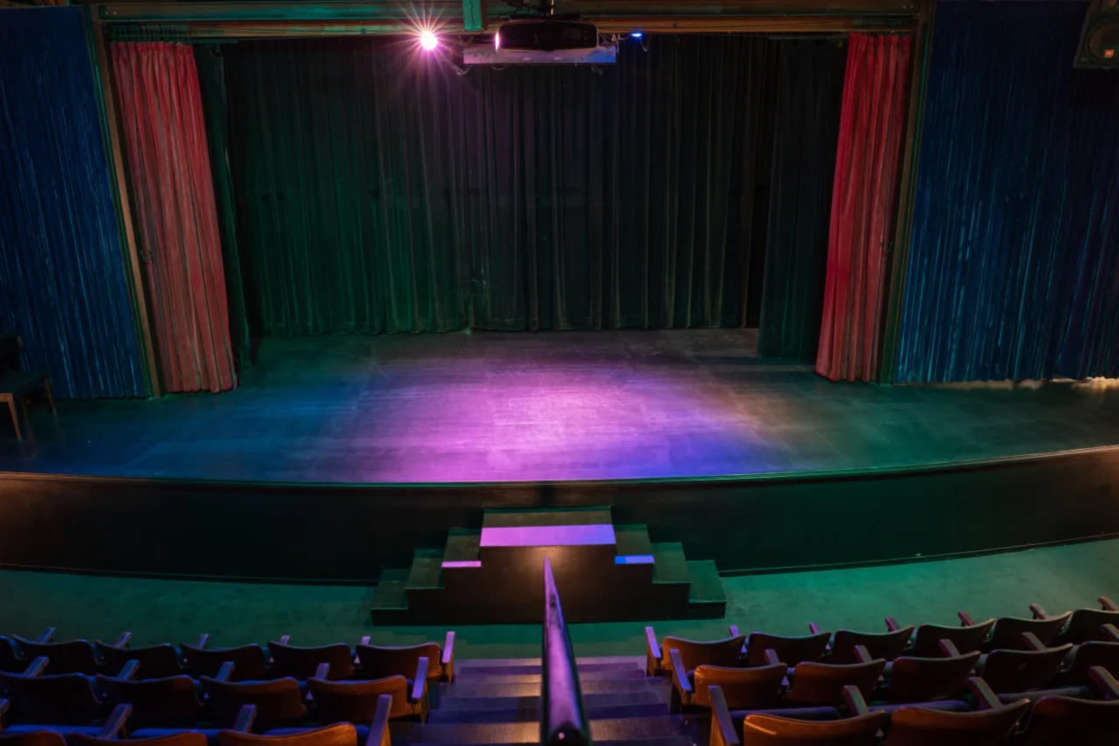 A theater stage with curtains and a seating area, illuminated by stage lights.