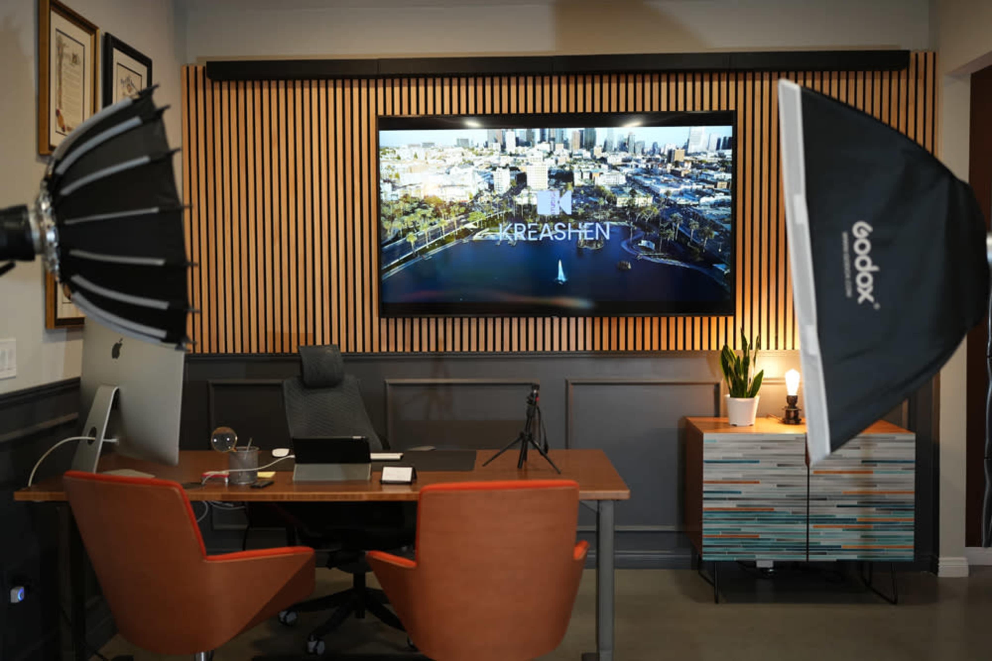 An office interior is shown with a large digital screen displaying a cityscape, a wooden wall feature, a desk with a chair, and studio lighting equipment placed nearby.