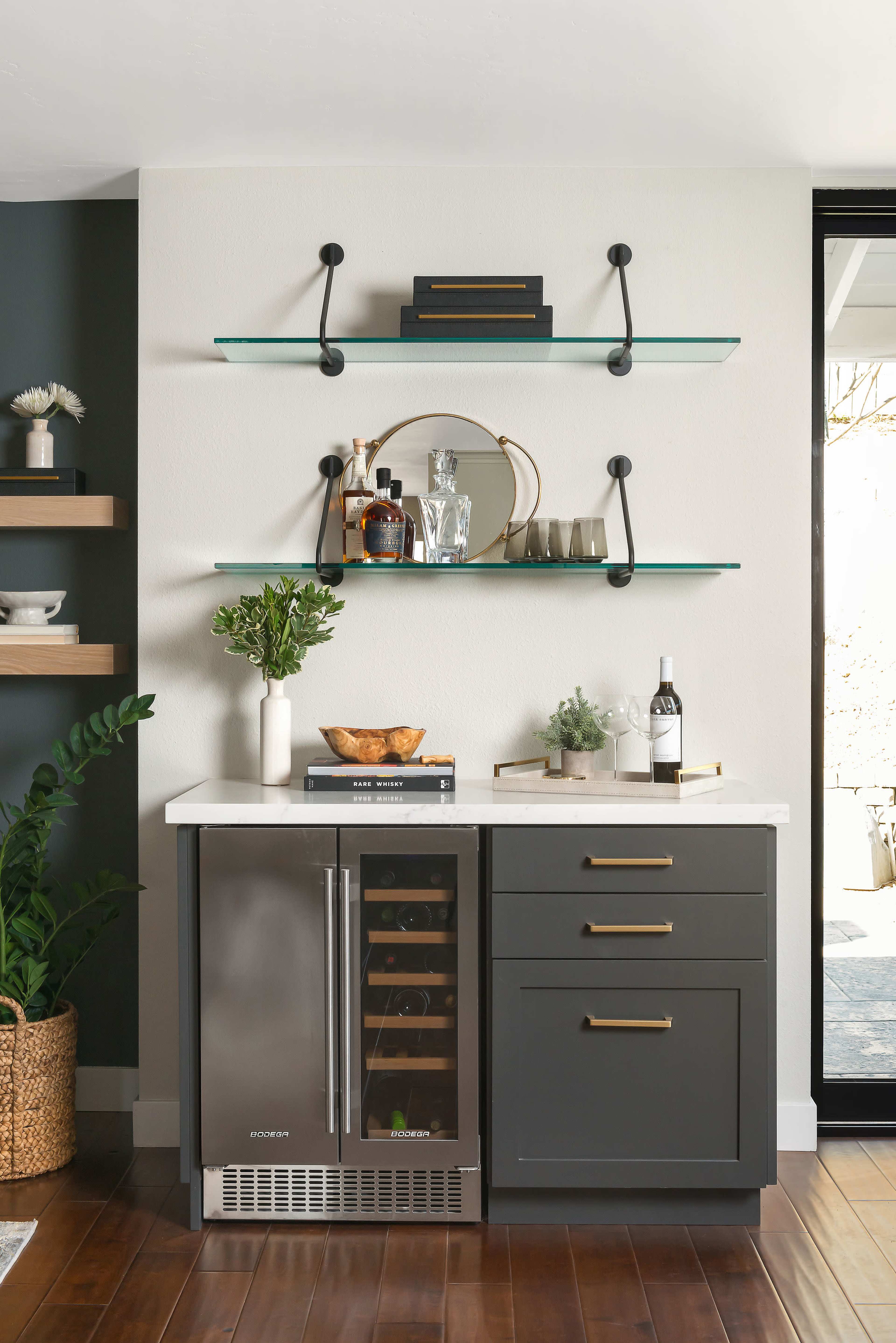 The image shows a modern kitchenette area featuring a wine cooler, storage cabinets, and open shelves with decorative items and glassware.