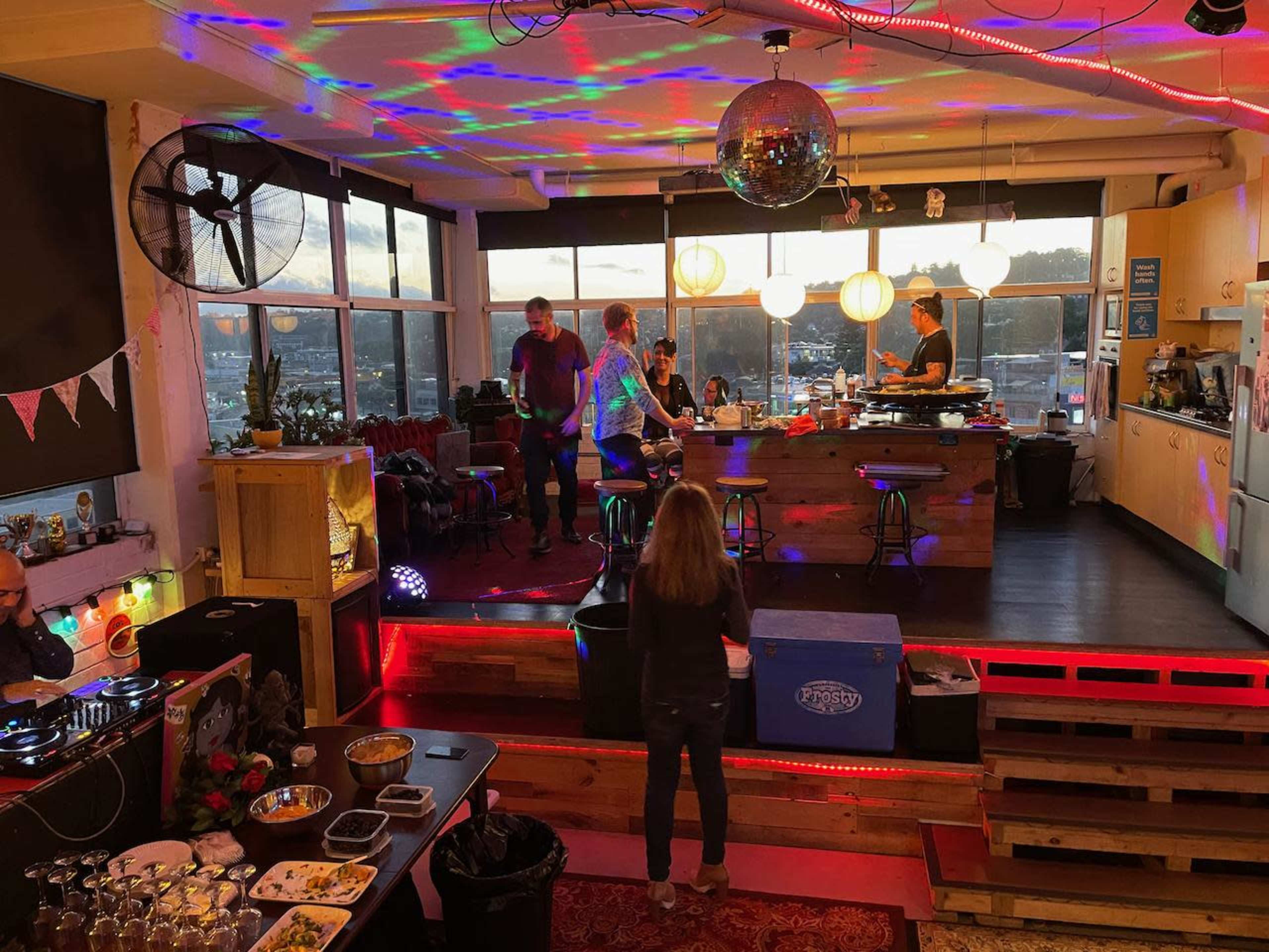 A group of people gathers in a brightly lit room with large windows, showcasing a sunset view, while preparing food and enjoying music amid colorful lights.