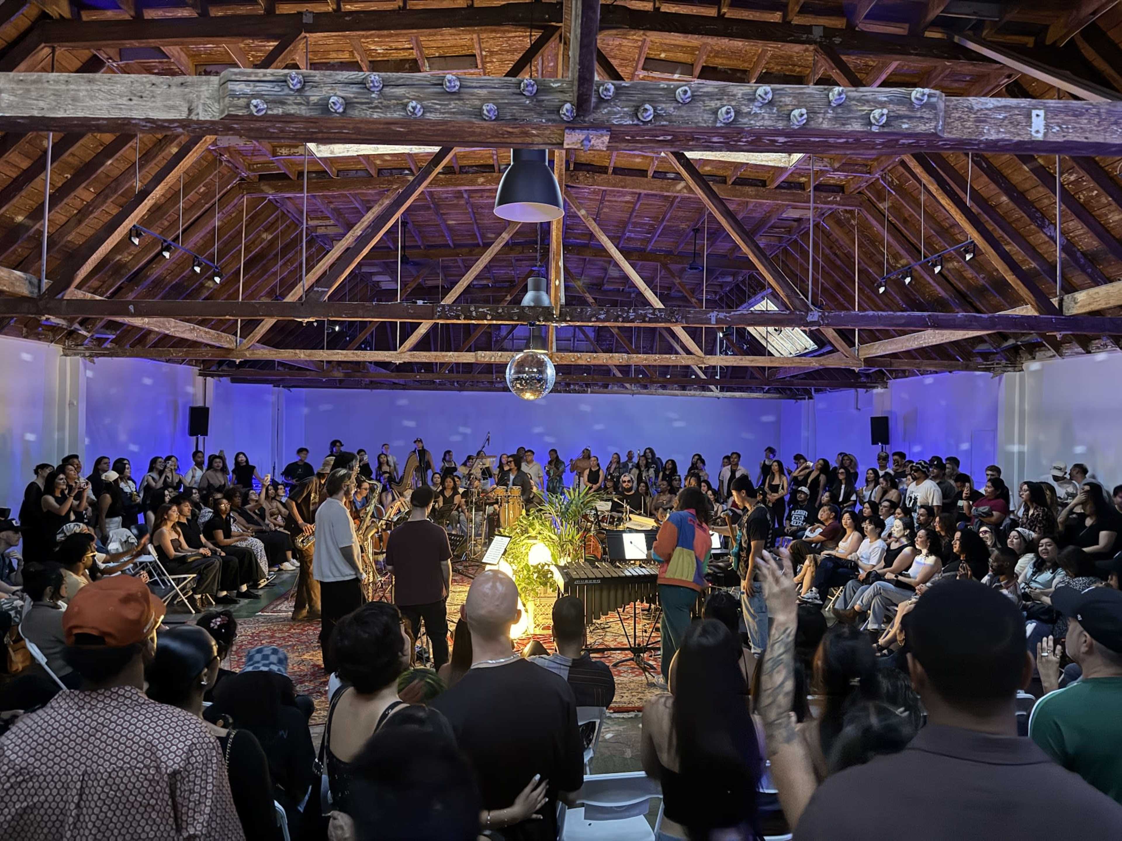 A large crowd gathers in a spacious venue with wooden beams and purple lighting, while performers play music in the center of the room.