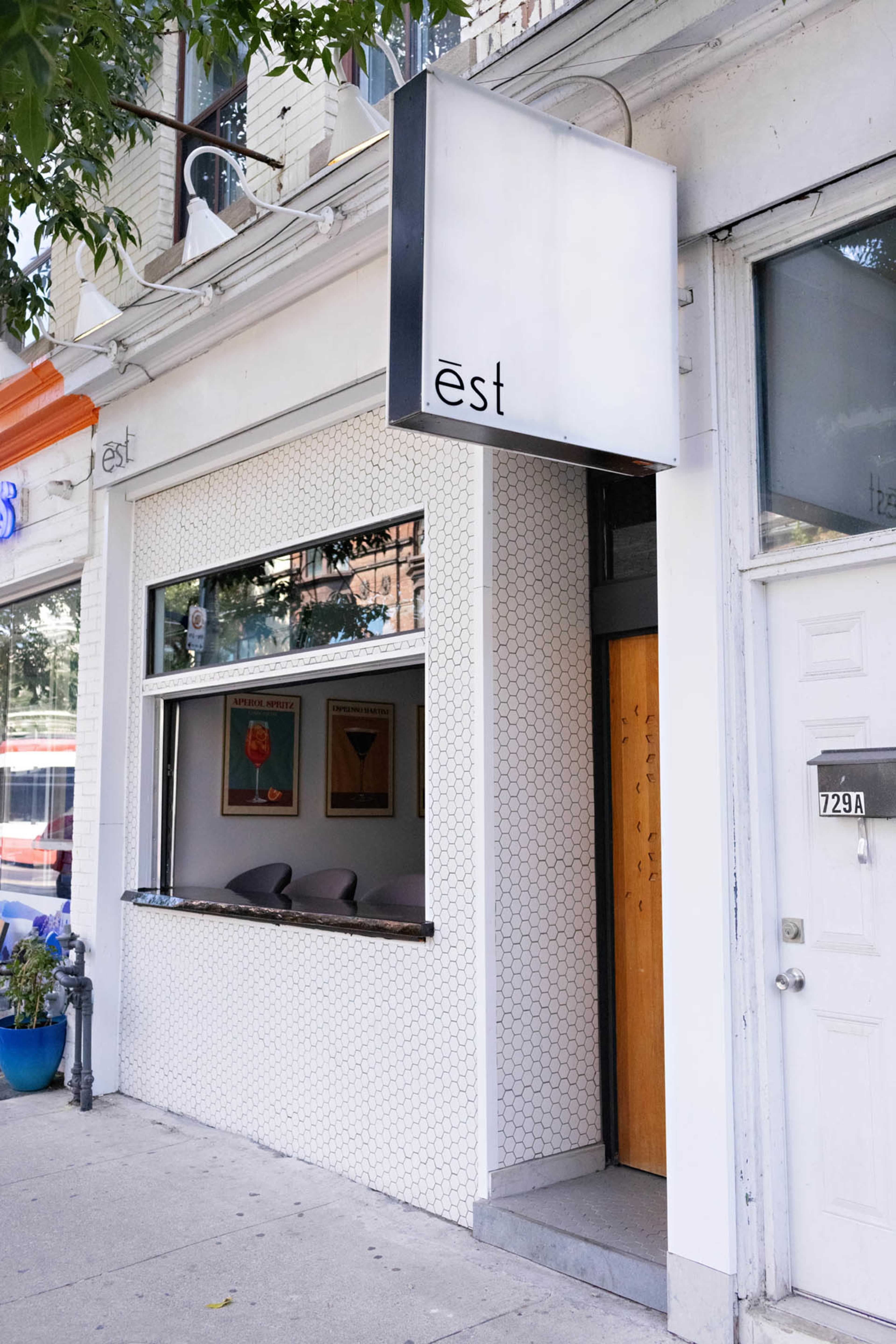 The exterior of a modern restaurant features a clean design with a large sign, a hexagonal tiled facade, and a window showing a seating area inside.
