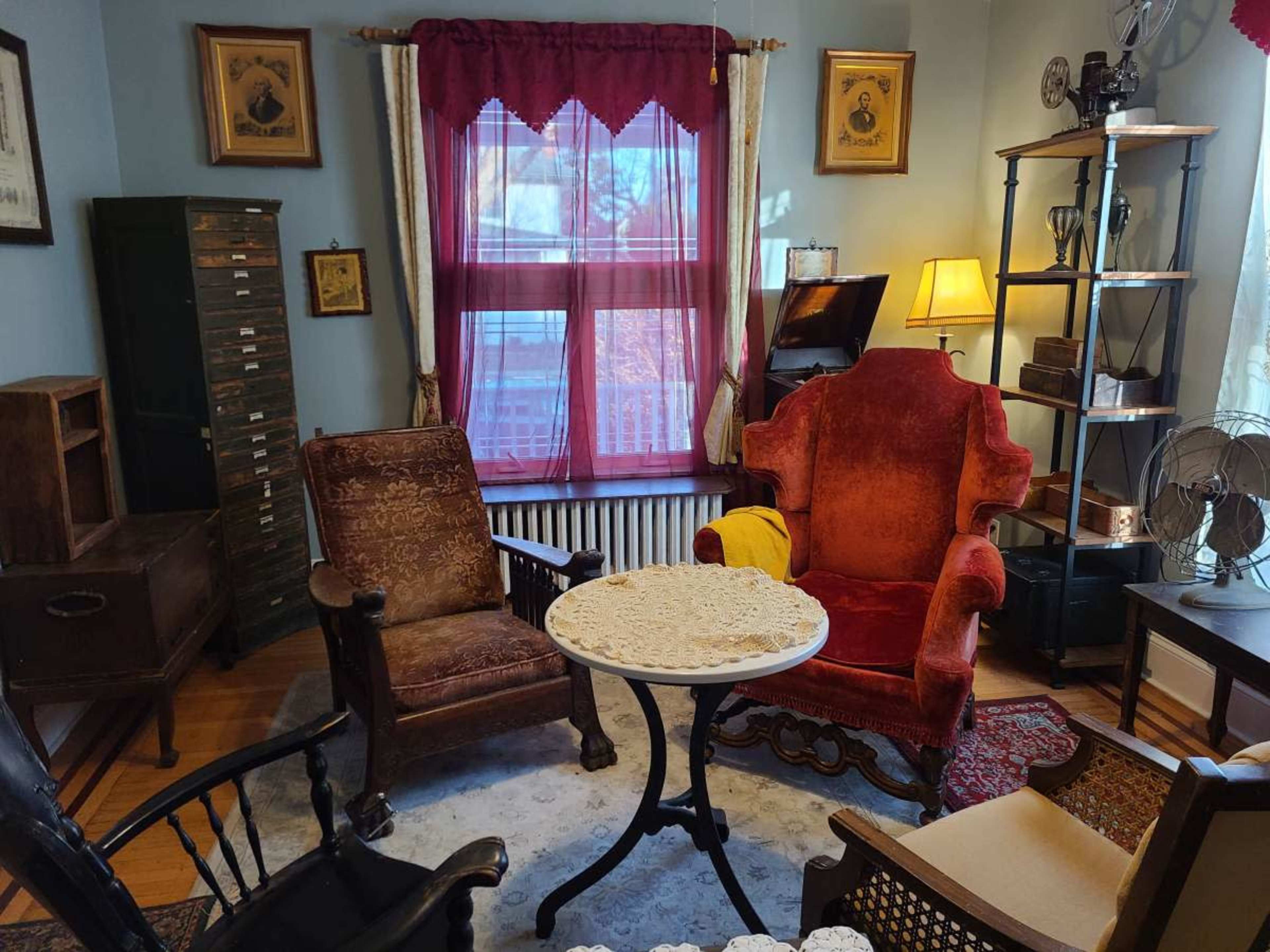 A cozy room furnished with a round table, two upholstered chairs, and various antique decorations, illuminated by natural light coming through a window.