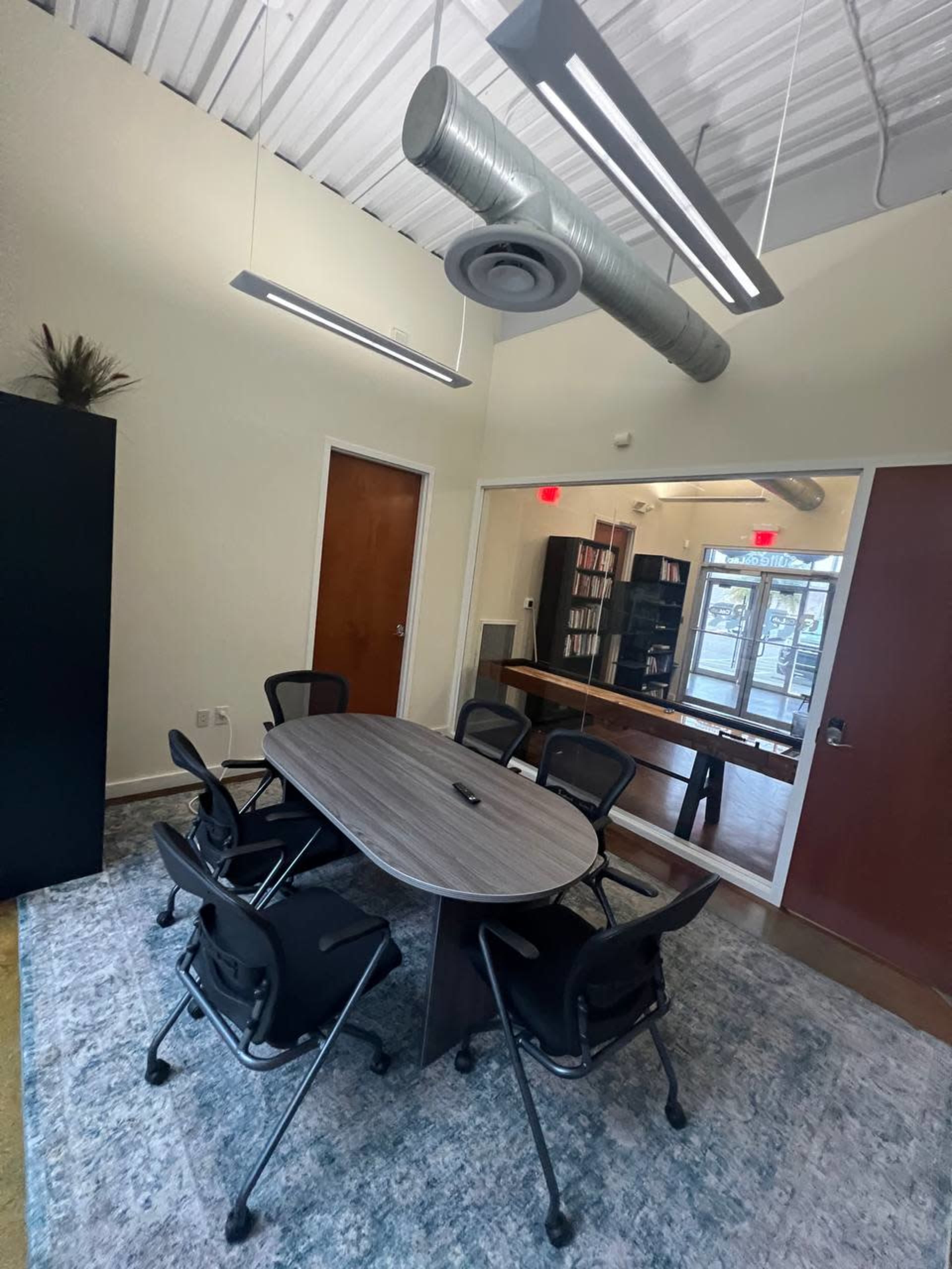 A modern conference room features a round table surrounded by six black chairs, with a view of a bookshelf and an adjoining room through a glass wall.