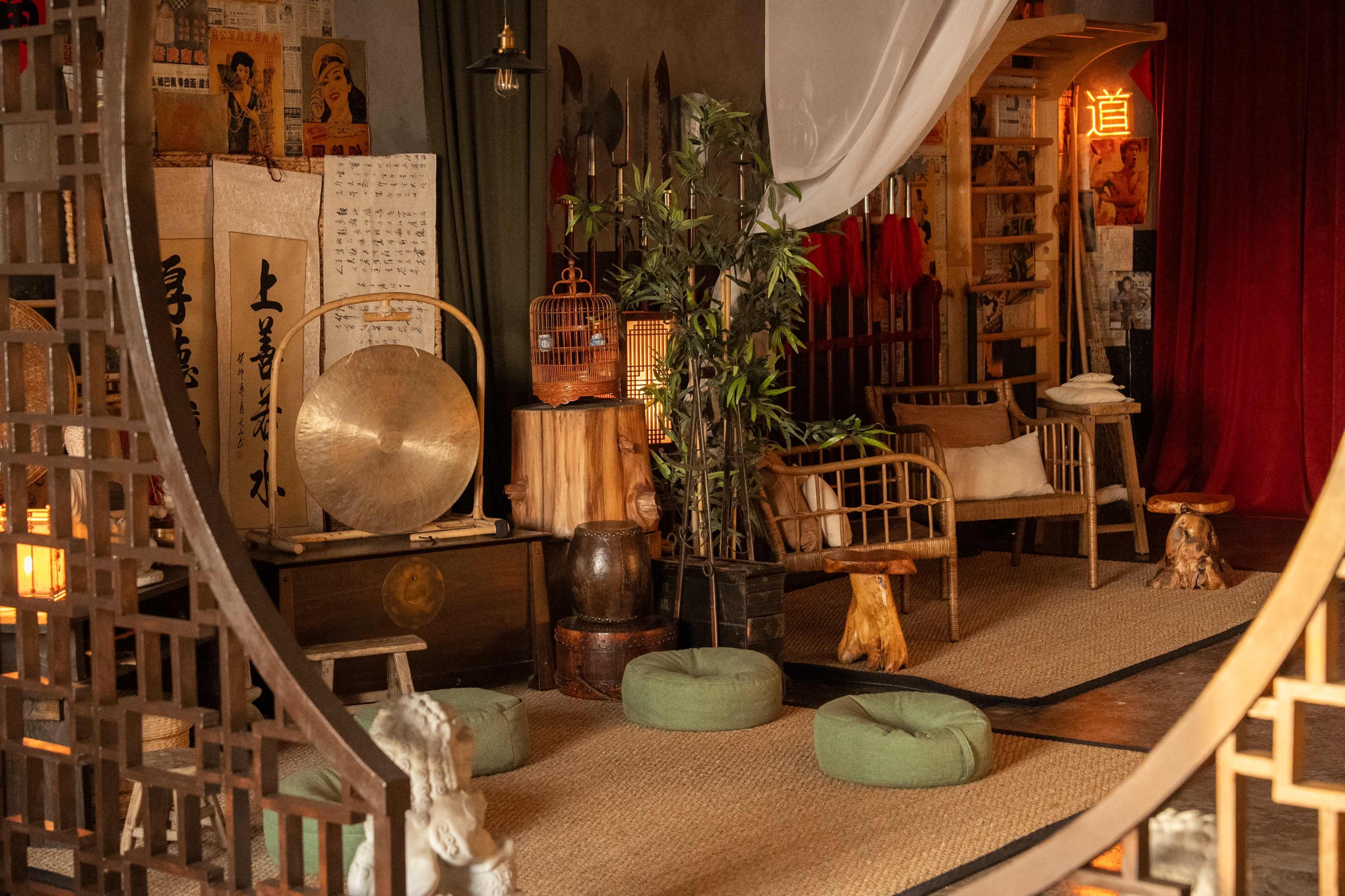 A cozy, eclectic interior space featuring bamboo furniture, a gong, and various decorative elements, including plants and soft seating arrangements.