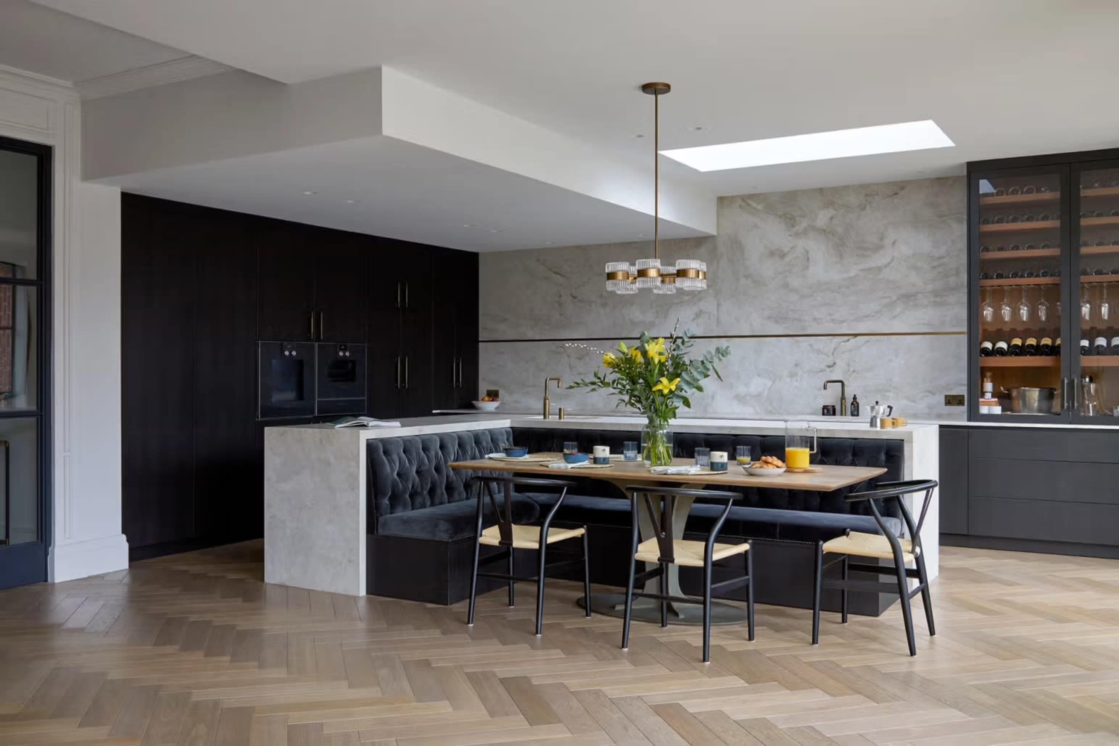 A modern kitchen features a large dining table surrounded by black chairs, with a plush banquette against the wall and light-colored cabinetry.