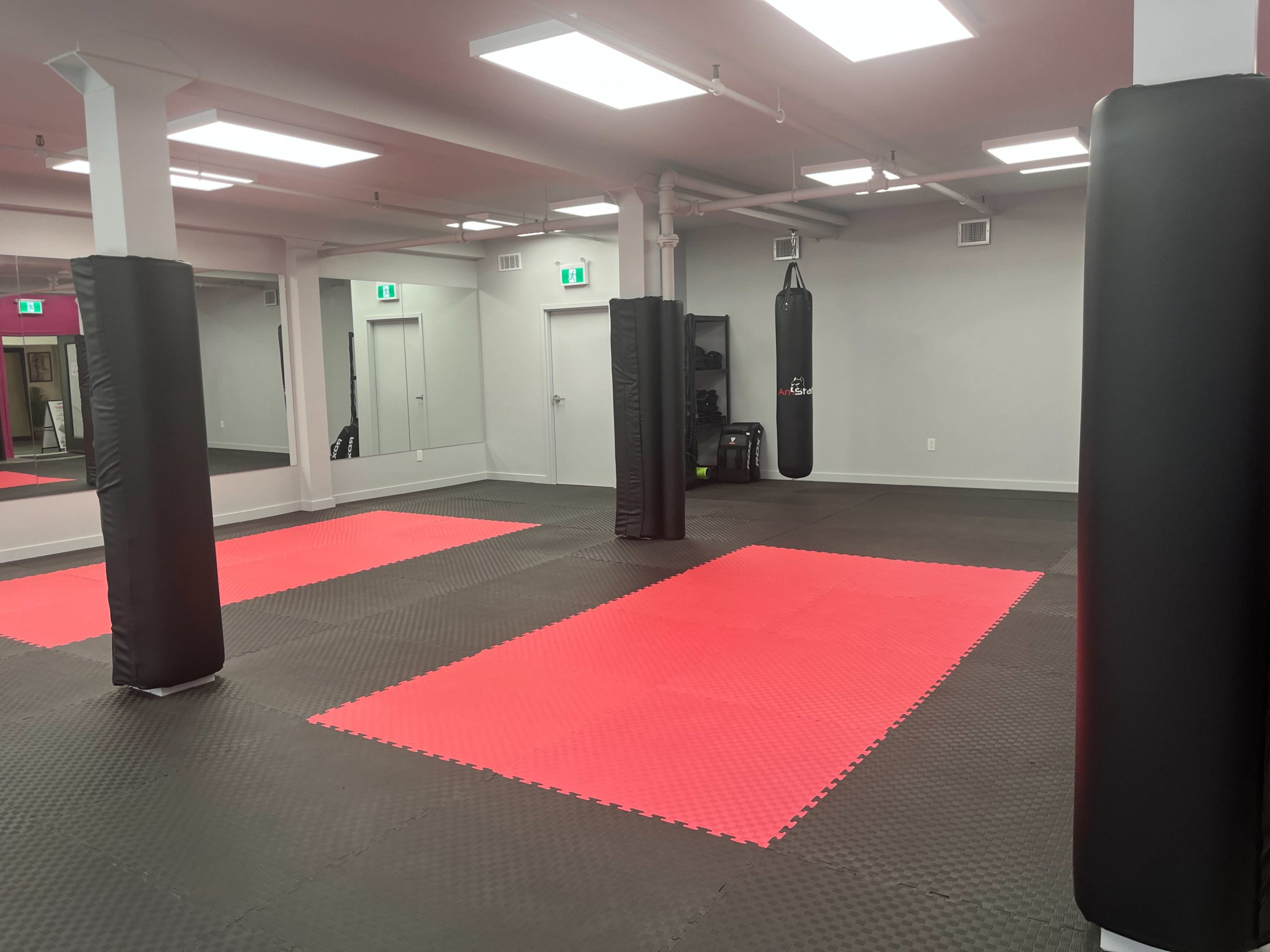 The image shows a spacious martial arts training studio with red and black flooring, black padded pillars, and a hanging punching bag.