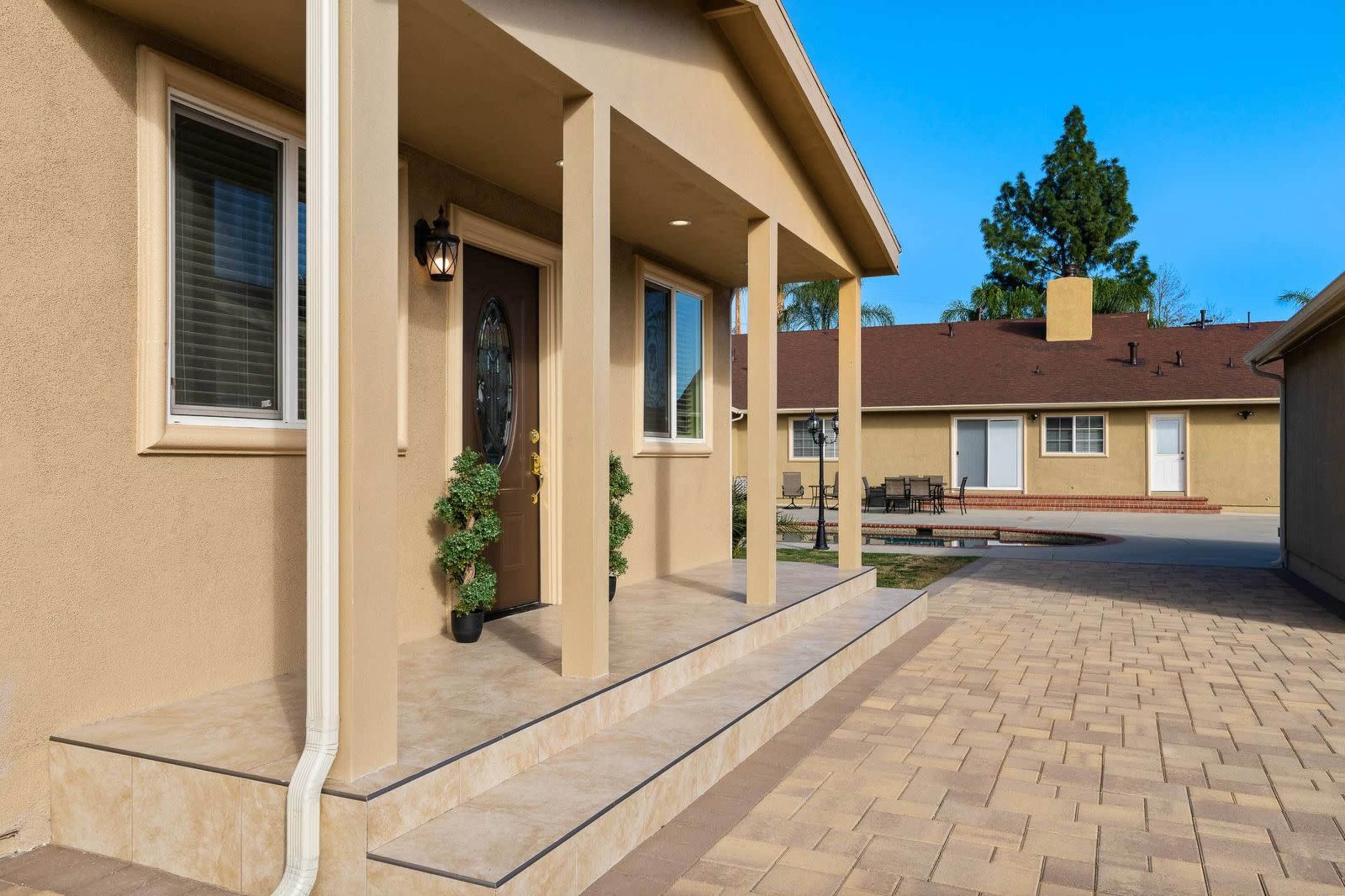 The image shows a house entrance with a covered porch, featuring a front door and potted plants, alongside a paved walkway leading to other buildings.