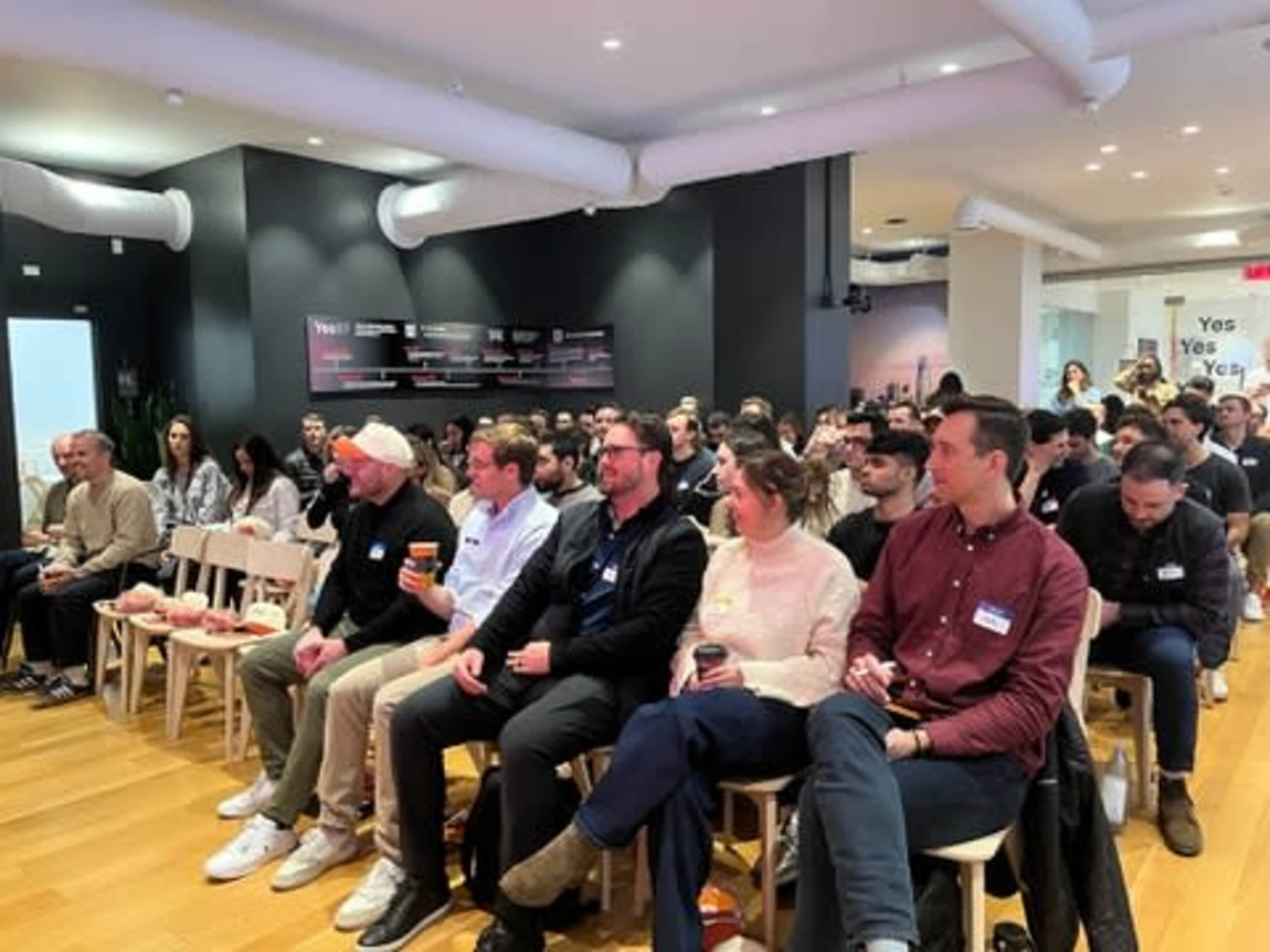 A crowd of attendees sits in rows at an event, facing a stage with a presentation in a well-lit indoor space.