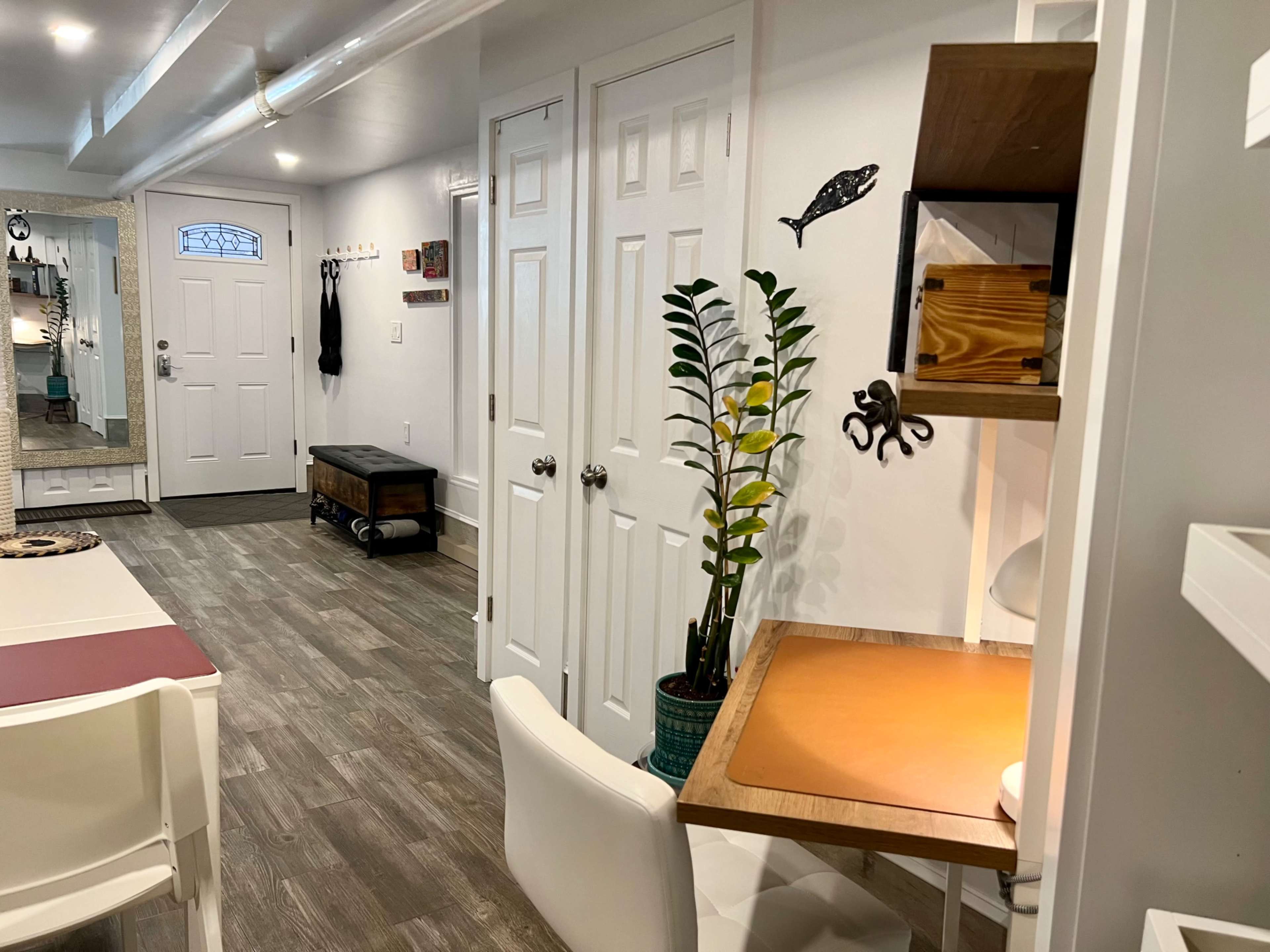 The image shows a well-lit interior space featuring a small desk with a chair, a plant, and white doors leading to a hallway and entry area.