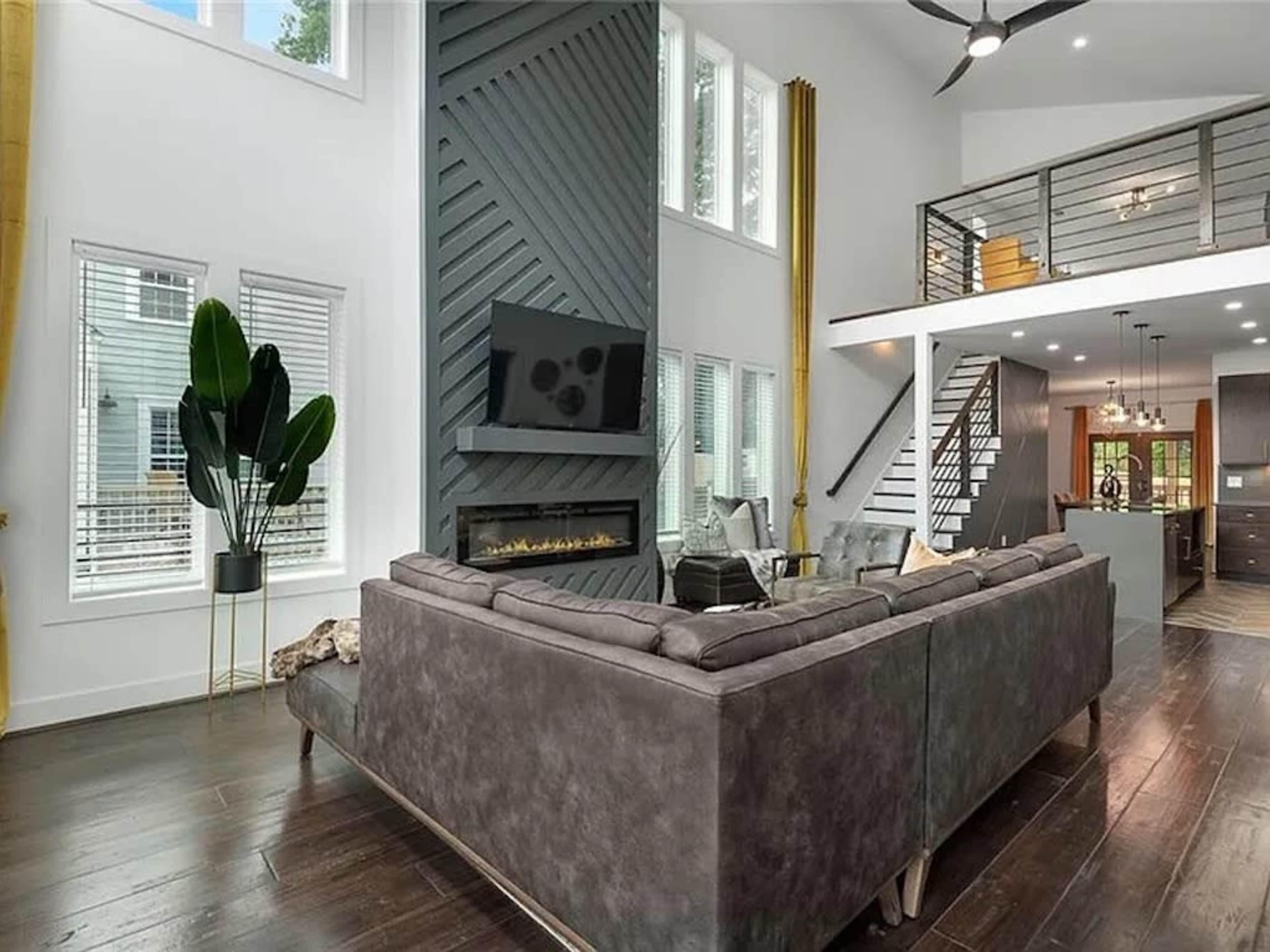 The image shows a modern living room with a sectional sofa, a vertical wood panel feature wall, a mounted television, and large windows allowing natural light.