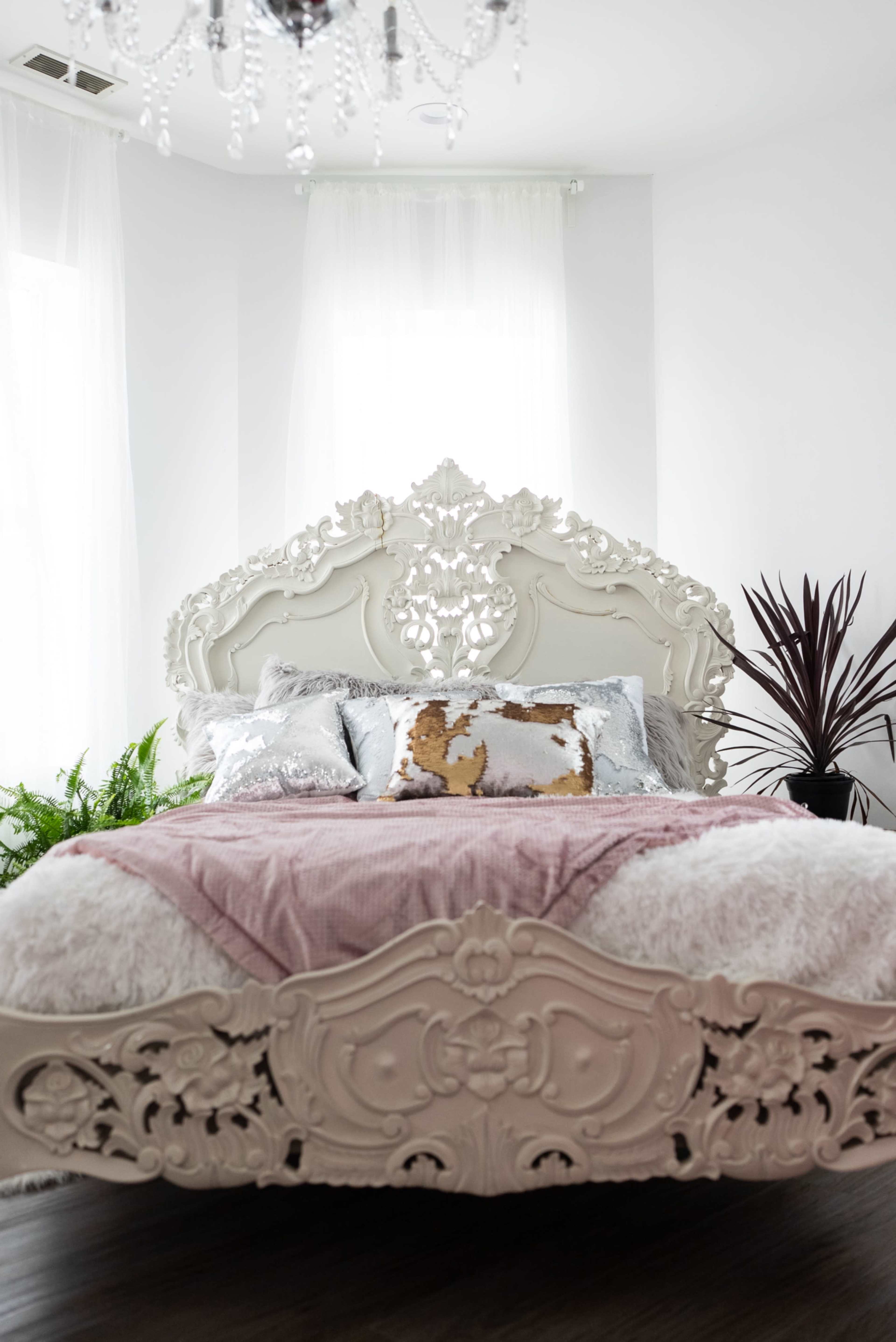 A decorative white bed with an ornate headboard is placed against a bright wall, surrounded by indoor plants and a chandelier.
