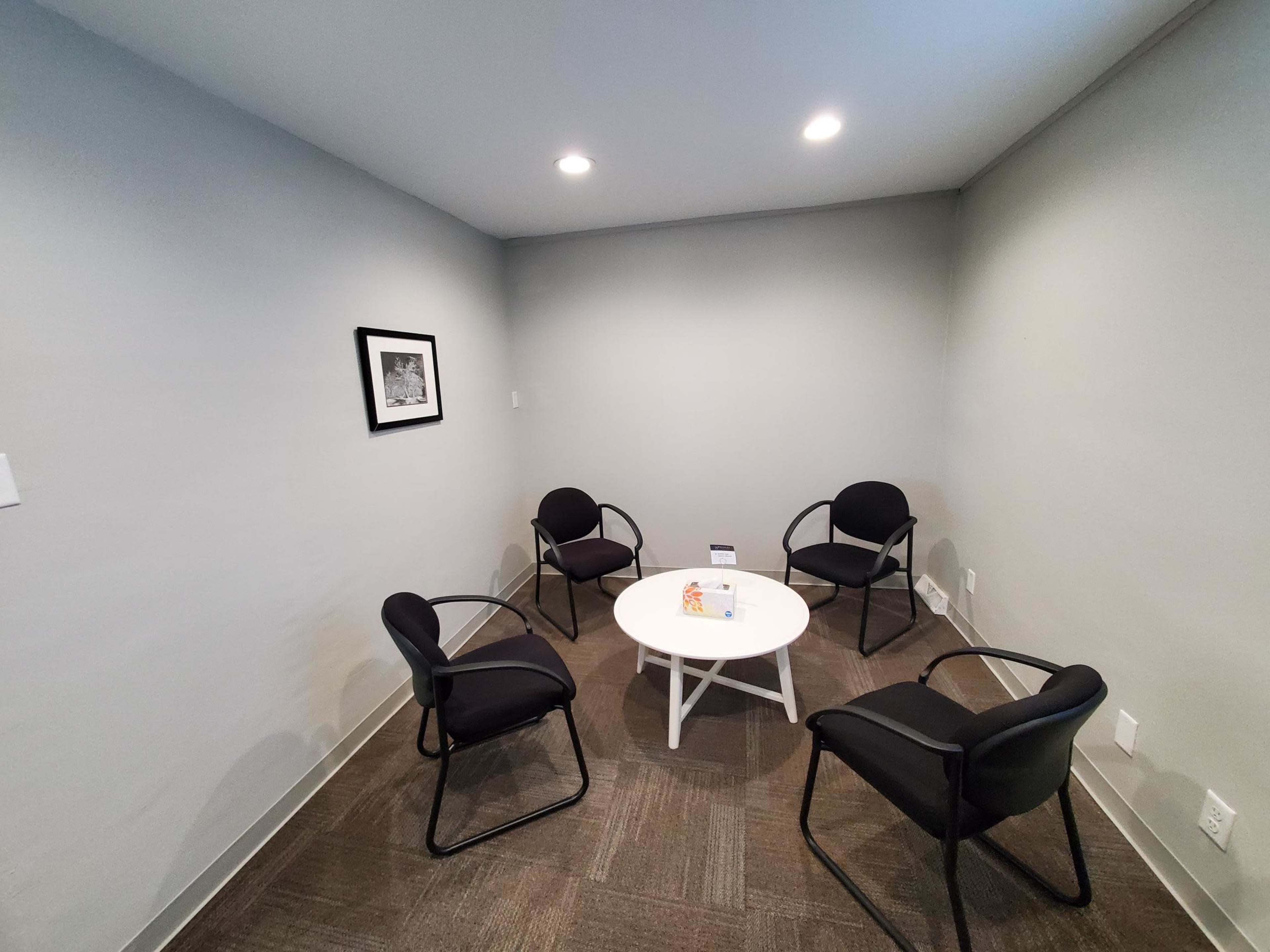 A small meeting room with four black chairs arranged around a white circular table on a gray carpet.
