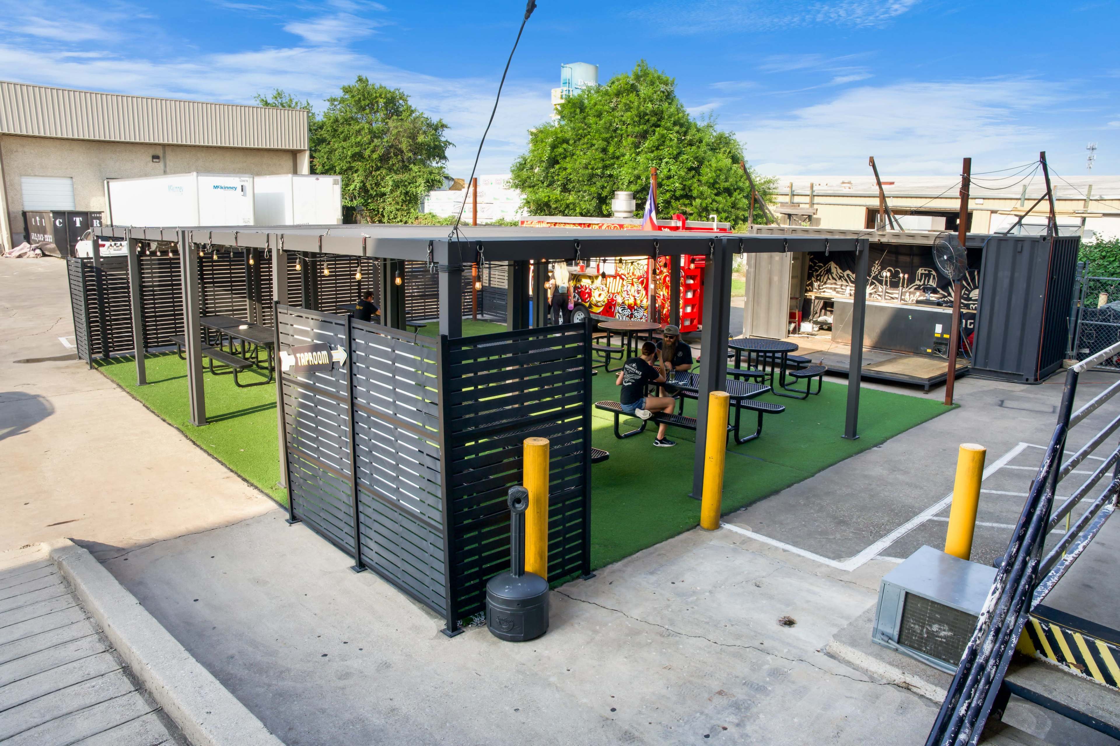 An outdoor seating area with picnic tables surrounded by black fencing, situated on a concrete surface with green grass and trees in the background.