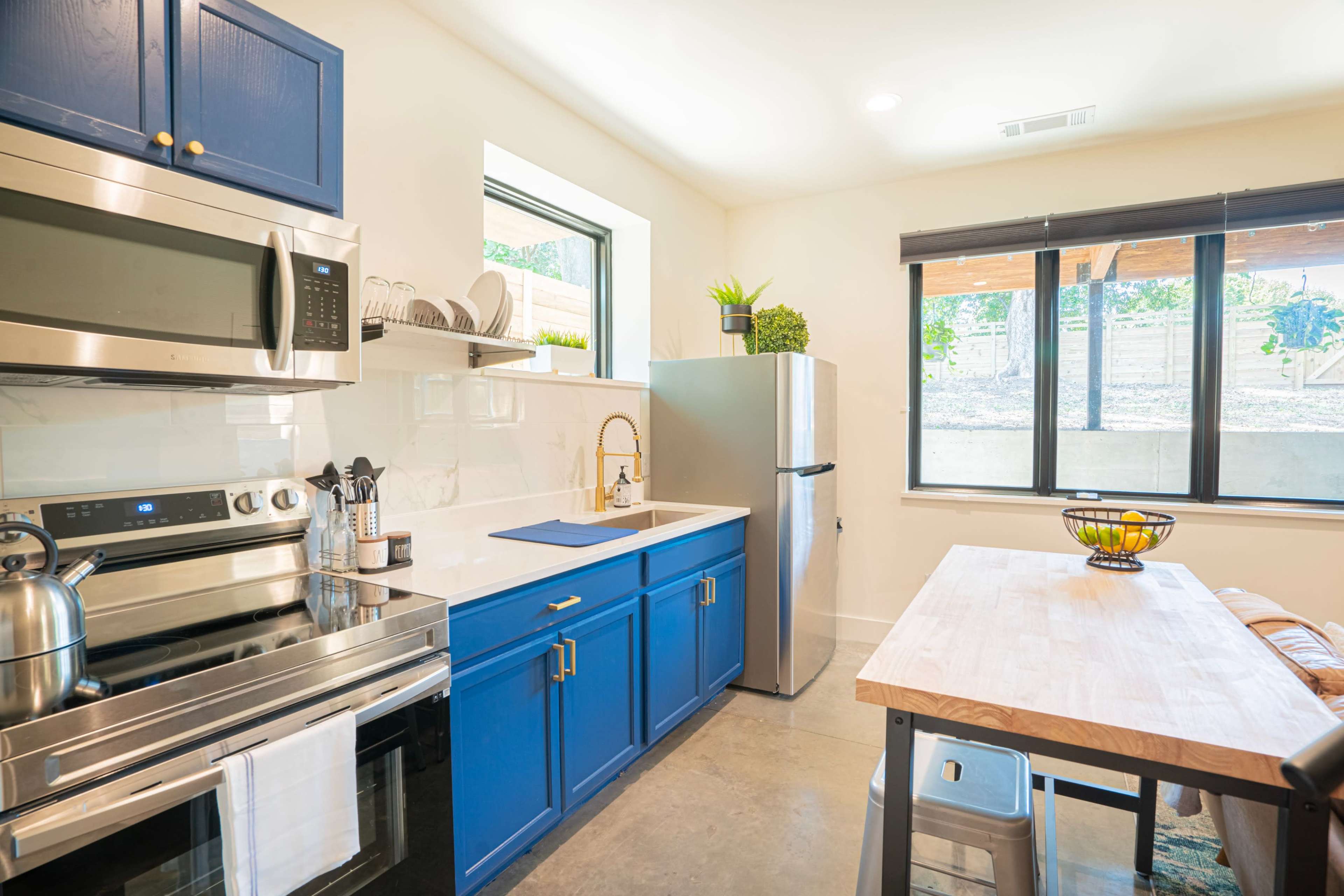 A modern kitchen features blue cabinets, stainless steel appliances, and a wooden dining table beside large windows.