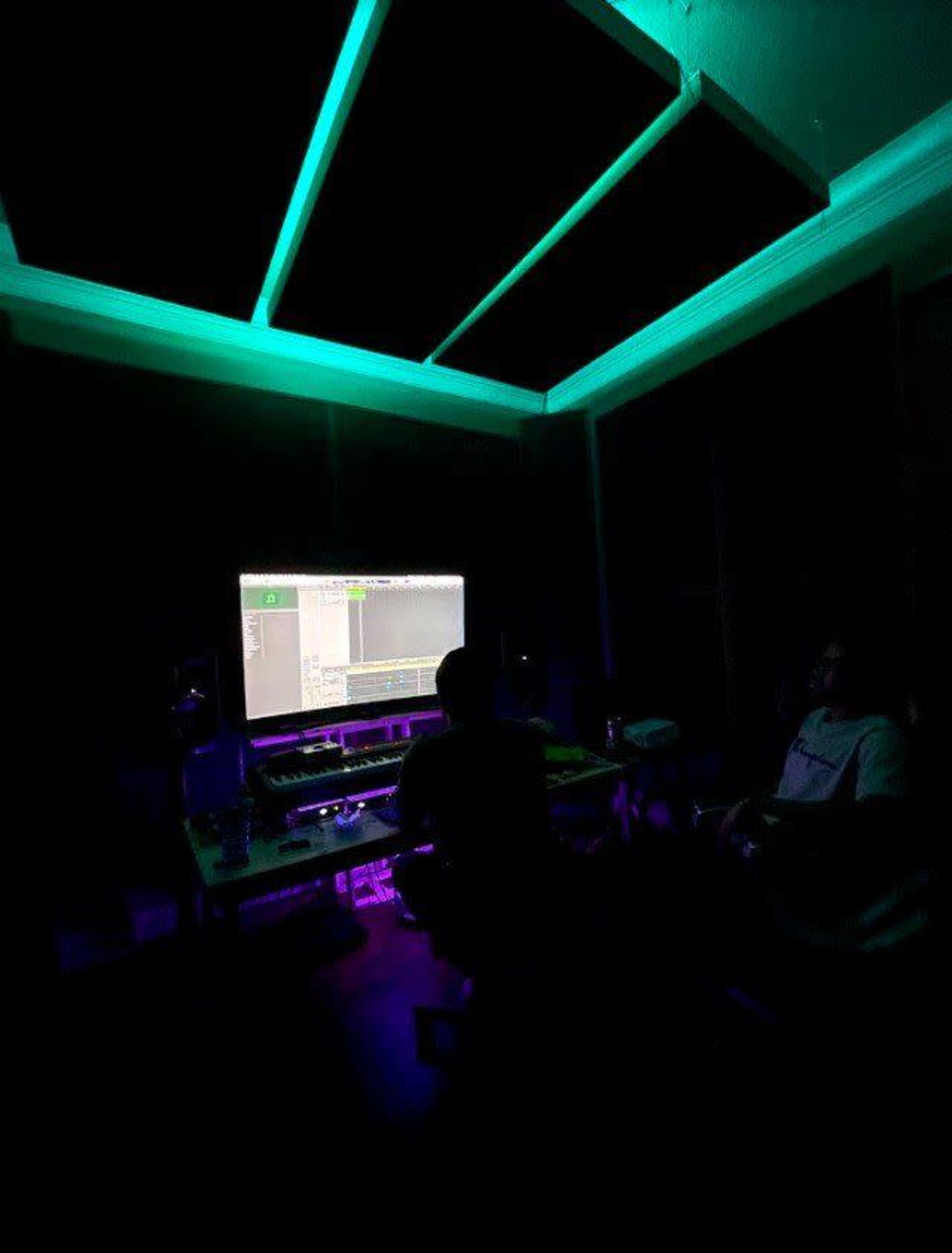 A person is sitting in a dimly lit music studio in front of a computer displaying audio editing software, with green and purple lighting highlighting the space.