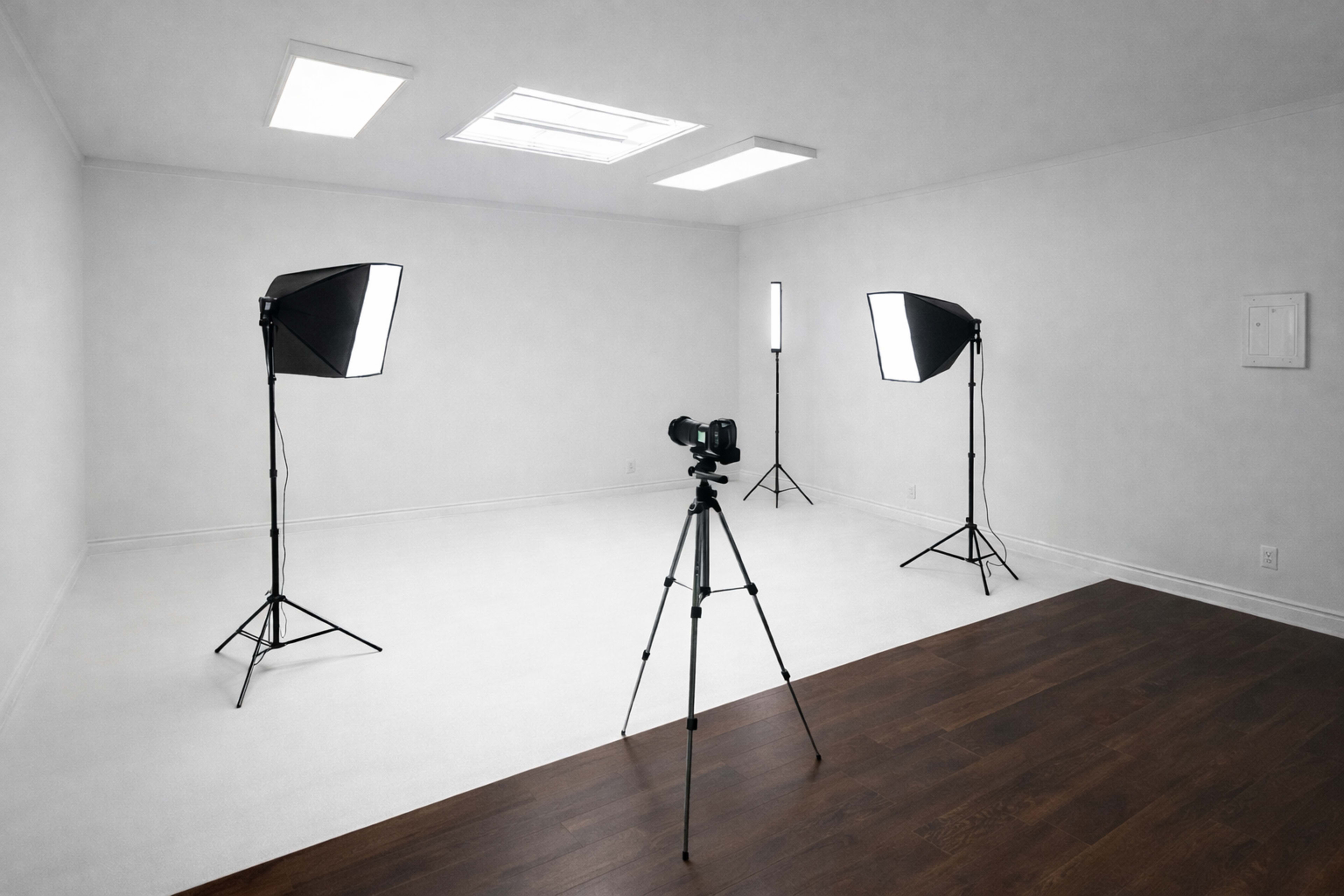 A photography studio features a camera on a tripod surrounded by two softbox light sources and an additional light in the corner.