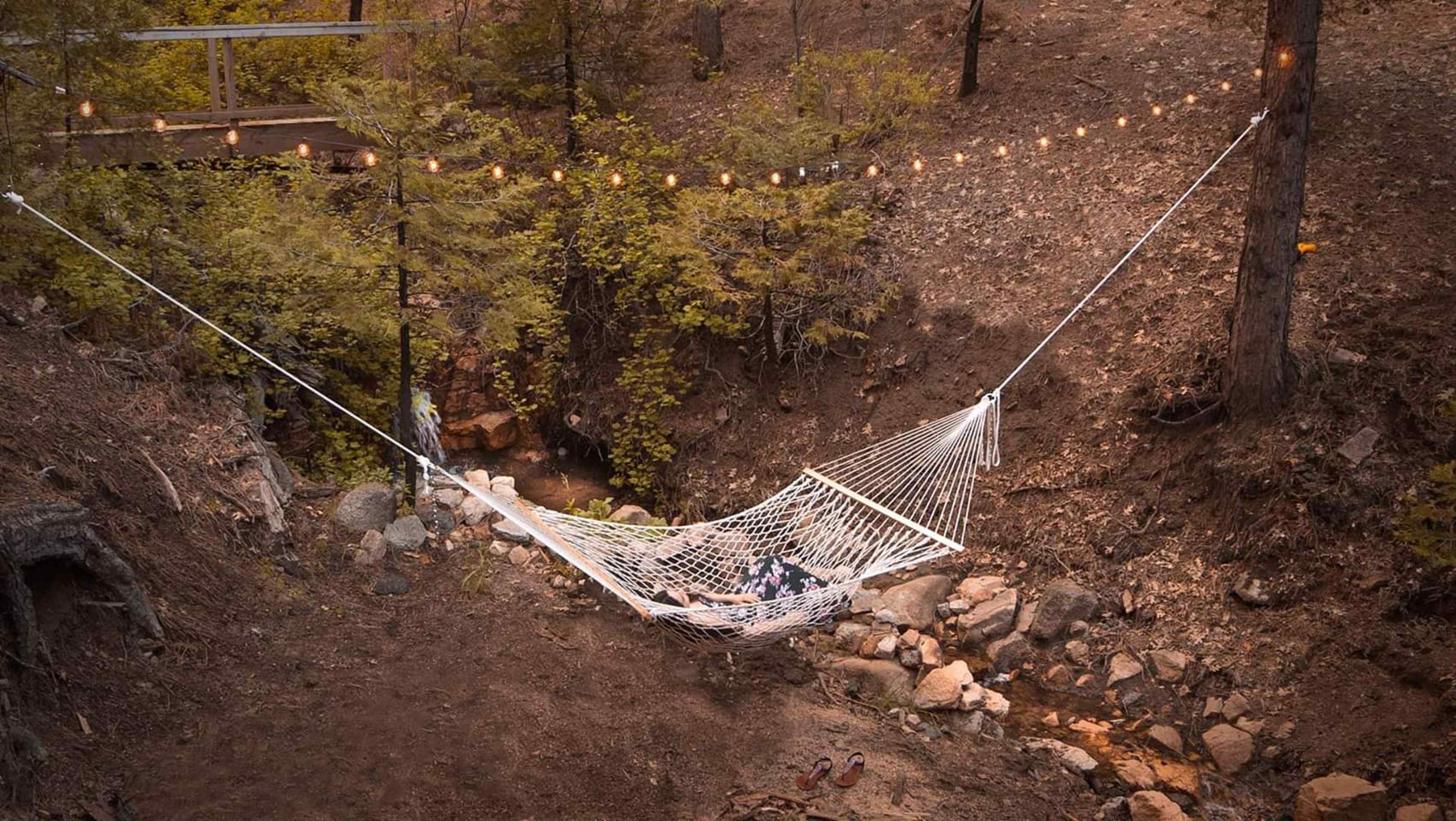 A white hammock is suspended between two trees above a rocky creek in a wooded area, with string lights hanging overhead.