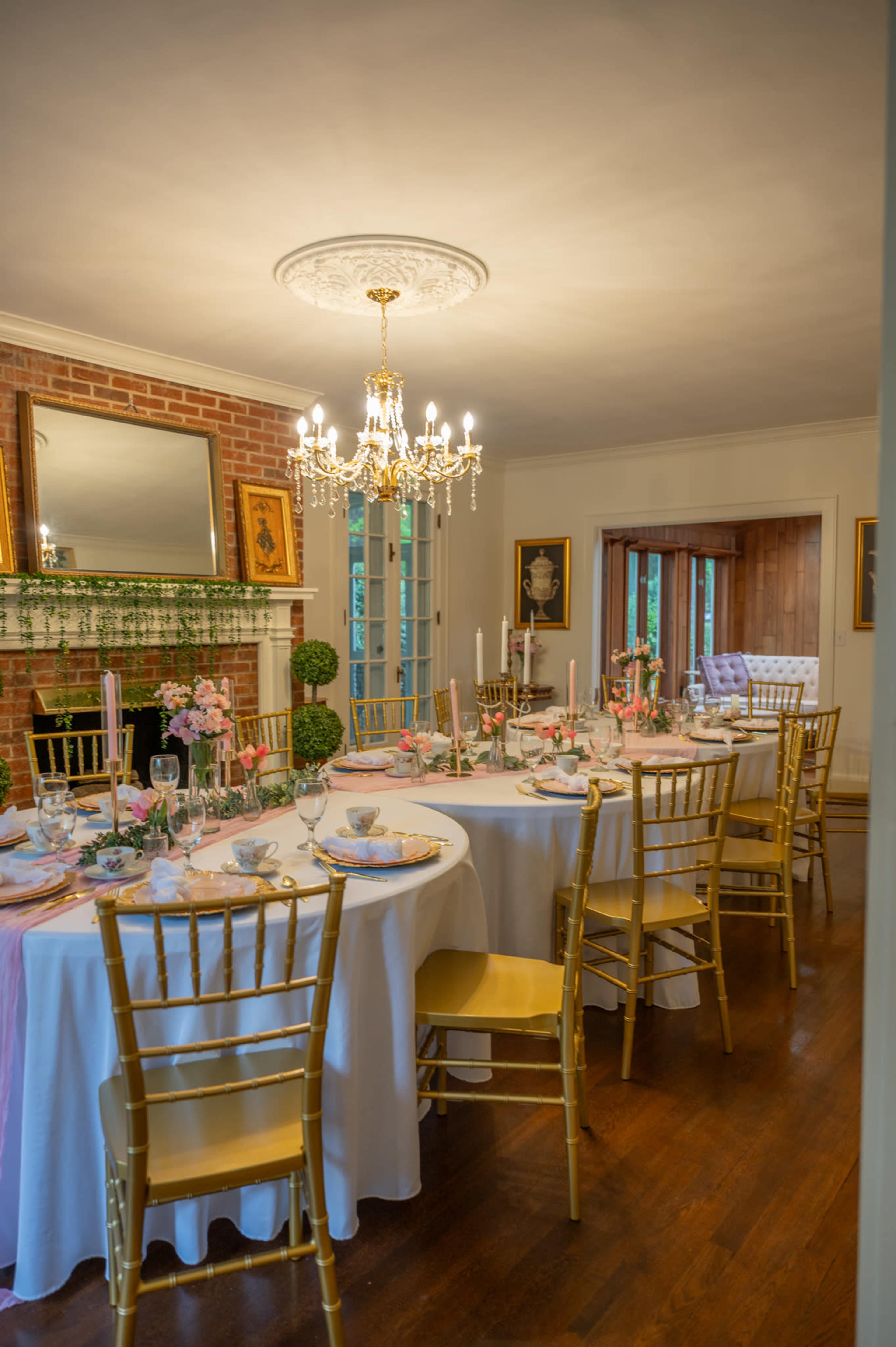 A dining room features a elegantly set table with golden chairs, a chandelier overhead, and a fireplace adorned with greenery and floral centerpieces.