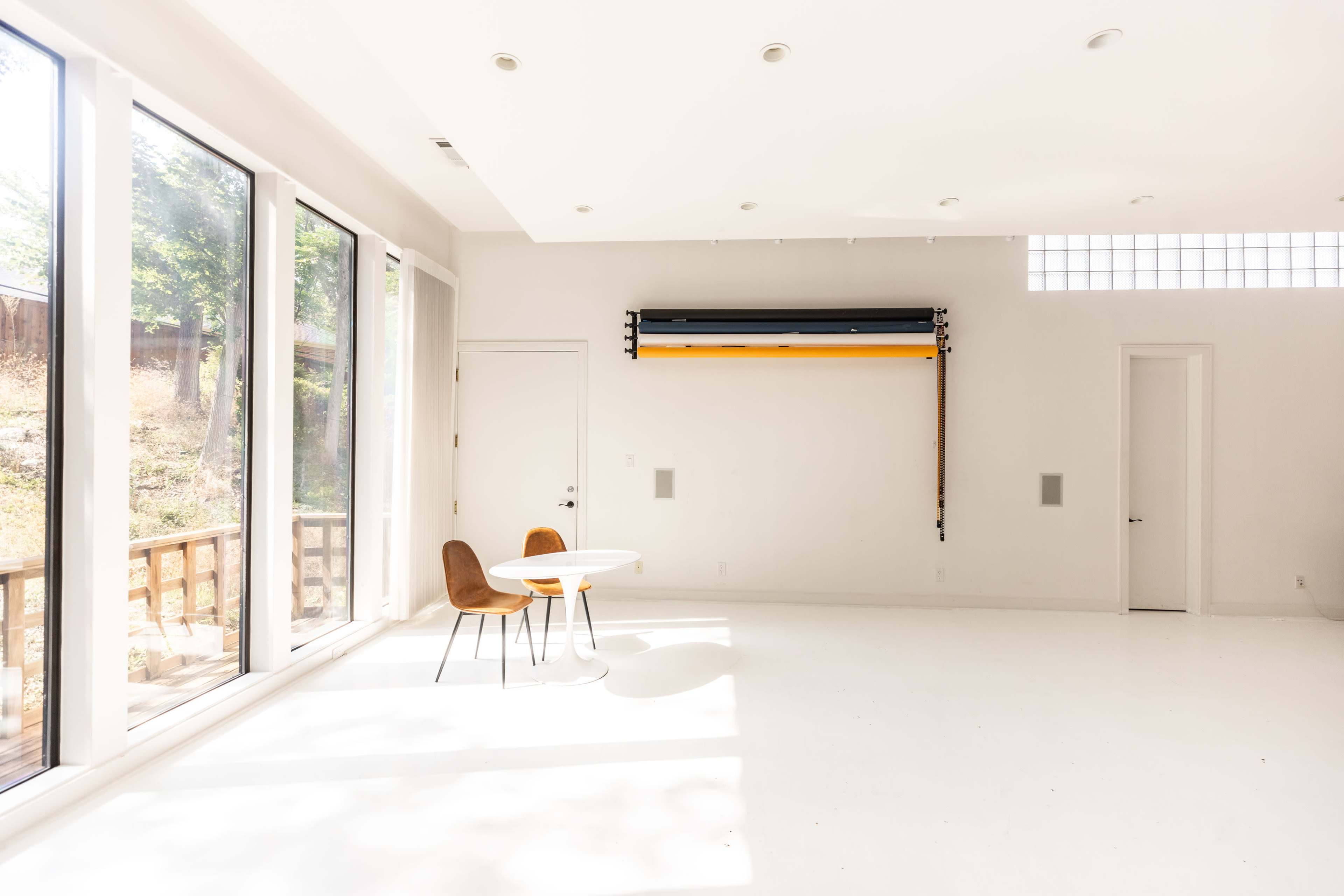 A bright, minimalist room features a round table with two chairs, large windows, and a roller shade mounted on the wall.