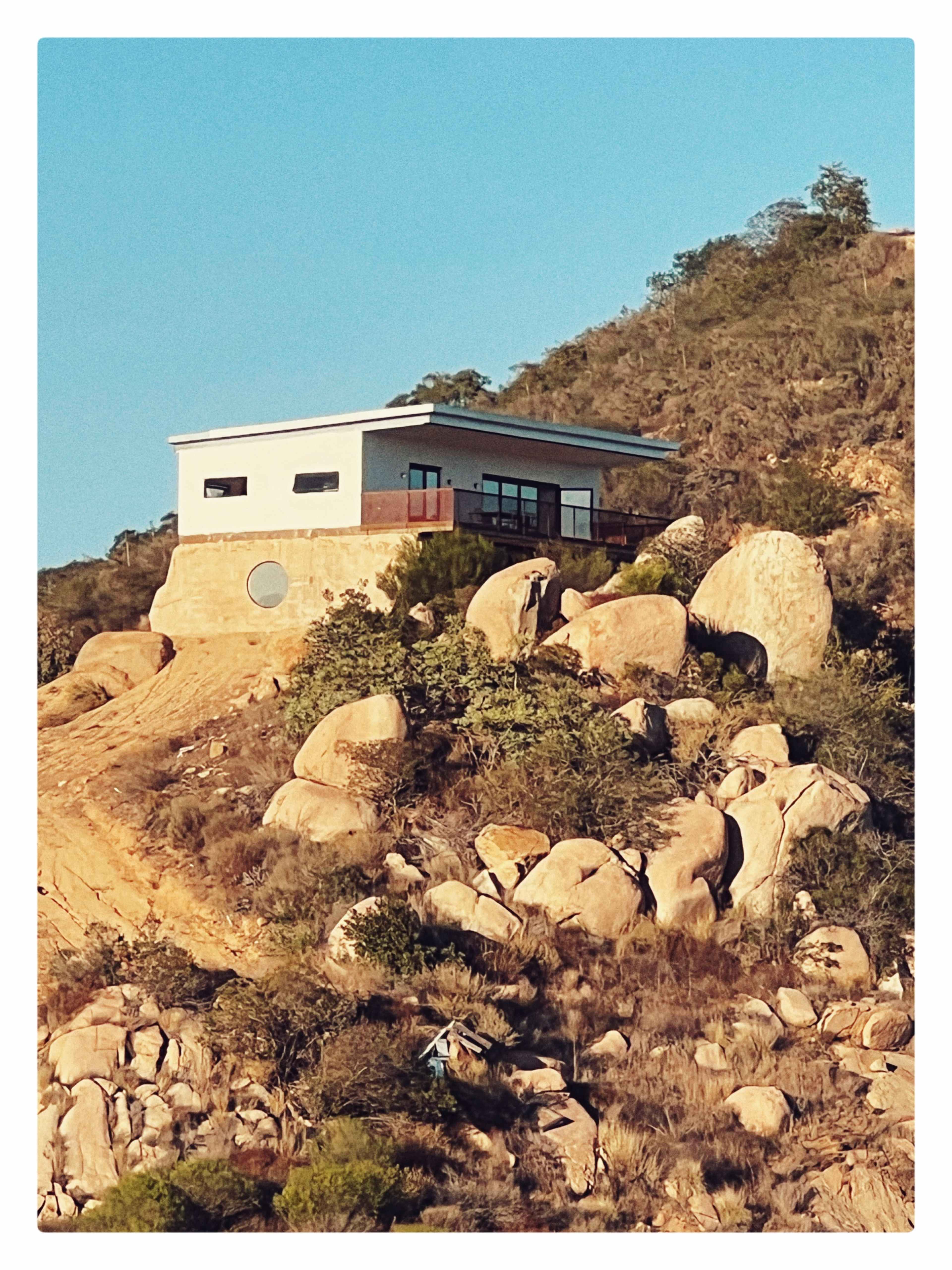 A modern house sits atop a rocky hill surrounded by large boulders and sparse vegetation.