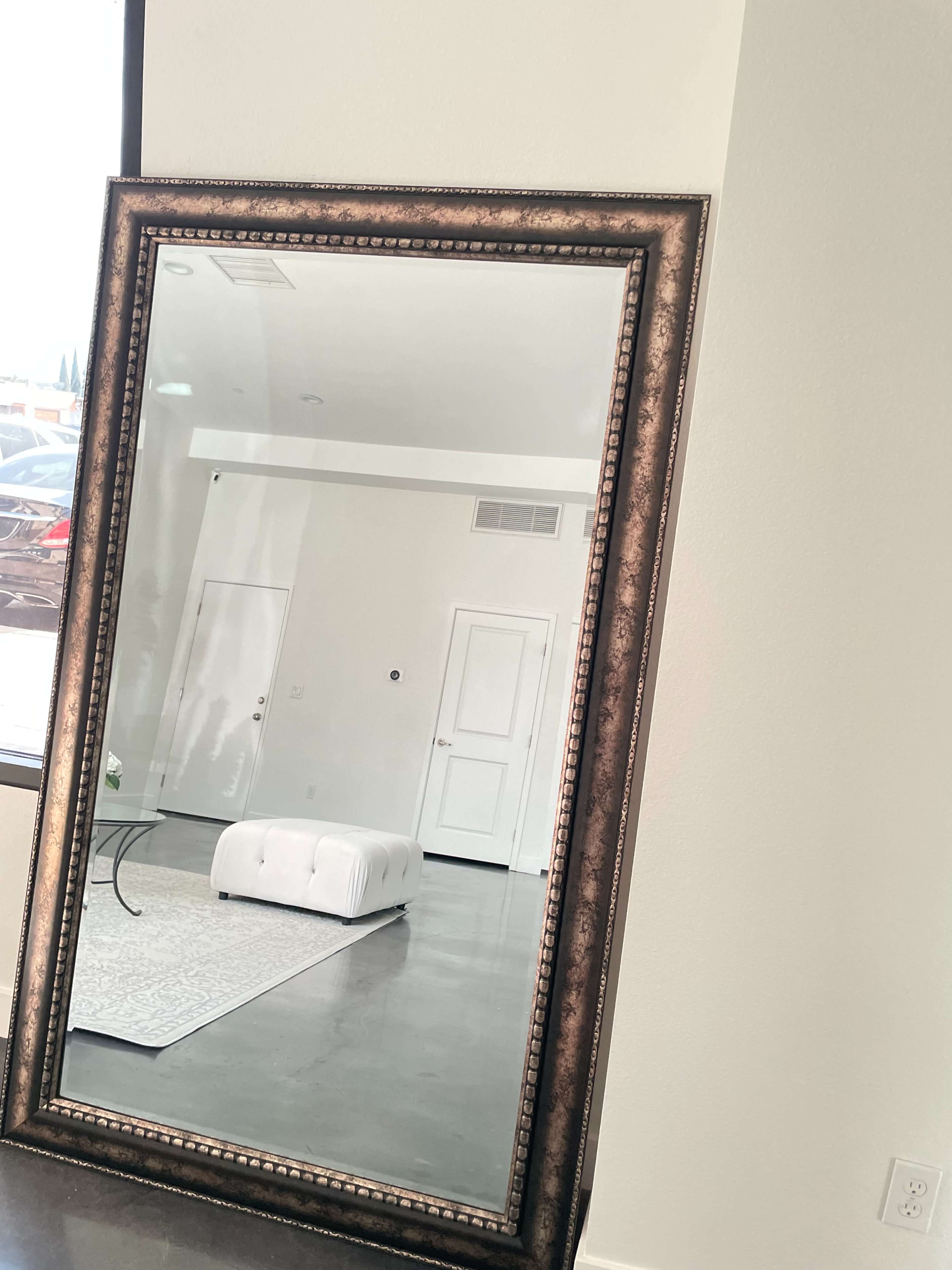 A large, ornate mirror is positioned against a light-colored wall, reflecting a minimalistic interior with a tufted white ottoman and a door in the background.