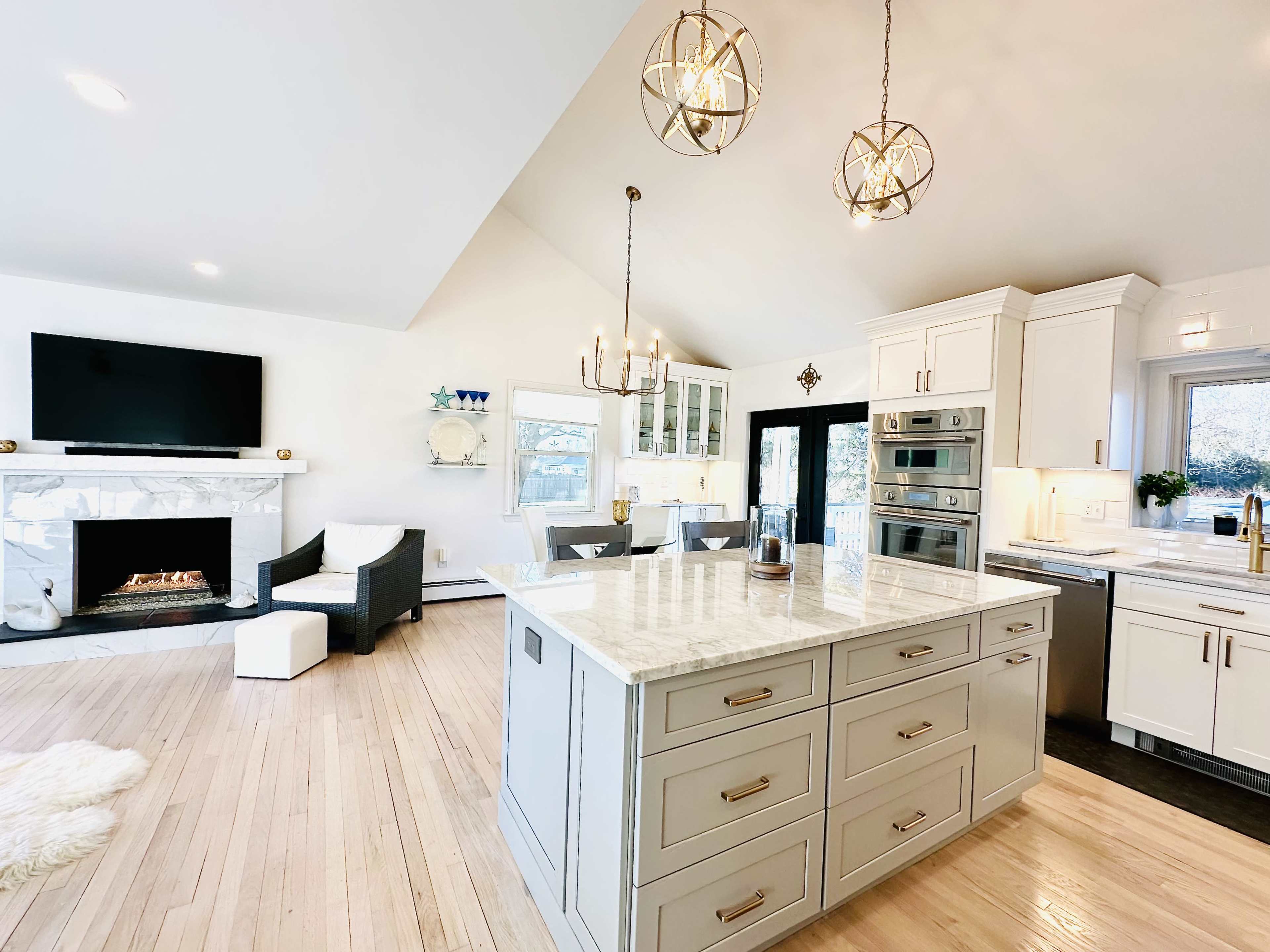 A modern kitchen with a large island featuring a marble countertop, surrounded by white cabinetry and a living area with a fireplace and a television mounted on the wall.