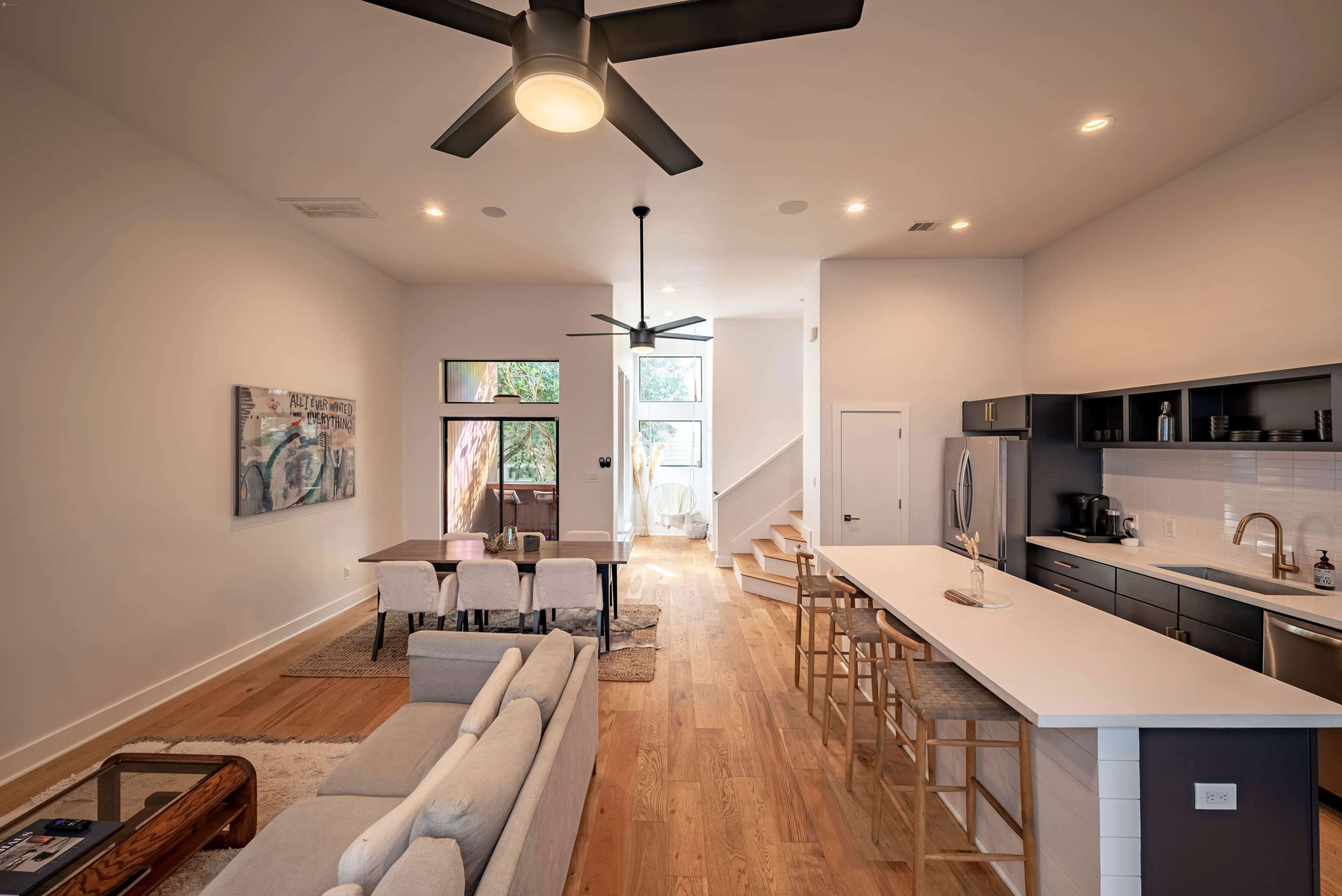 The image shows a modern open-concept living space with a kitchen island, dining area, and seating area, illuminated by natural light from large windows.
