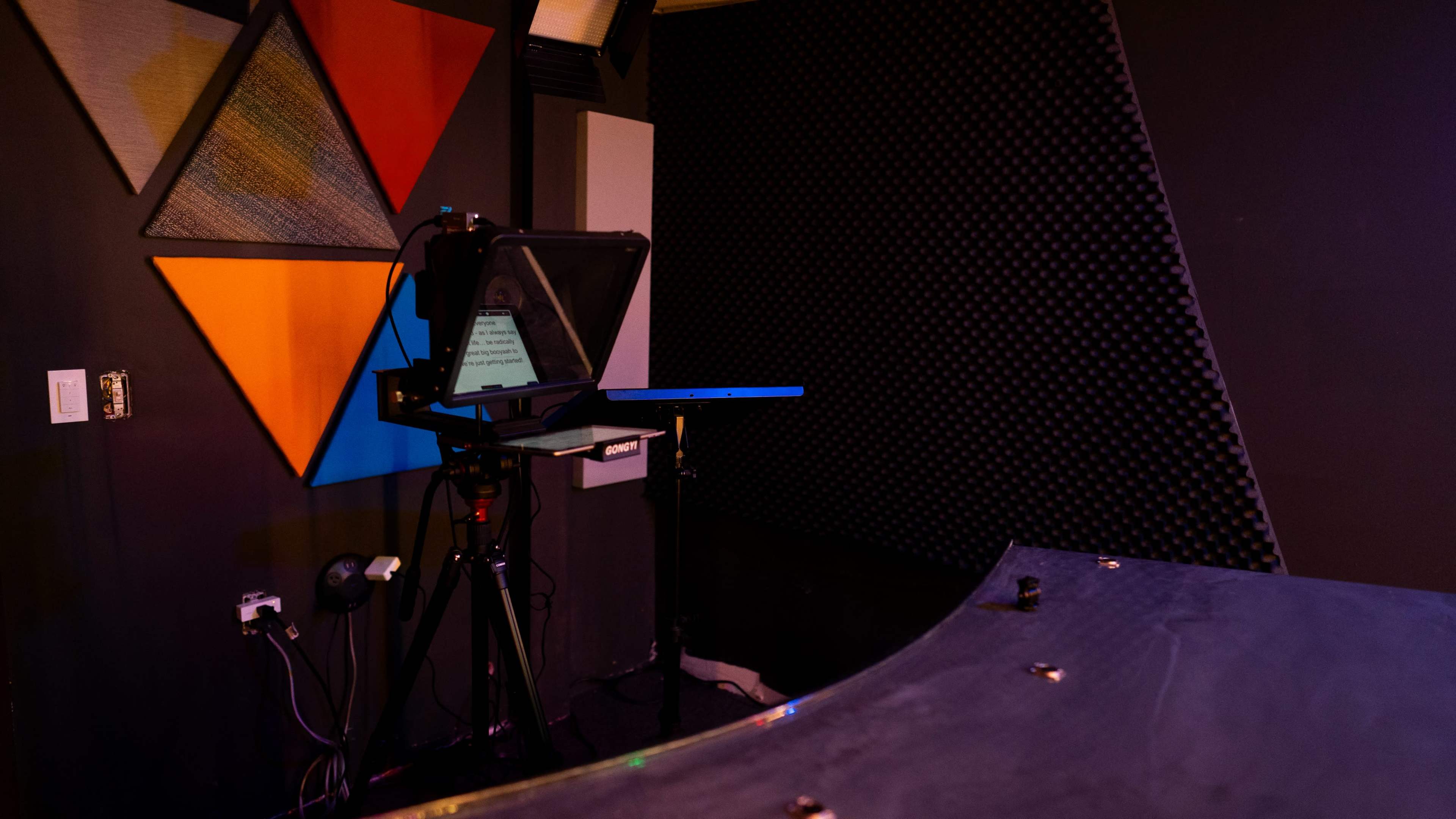A teleprompter is set up on a stand in front of a colorful geometric wall and a textured soundproof background.